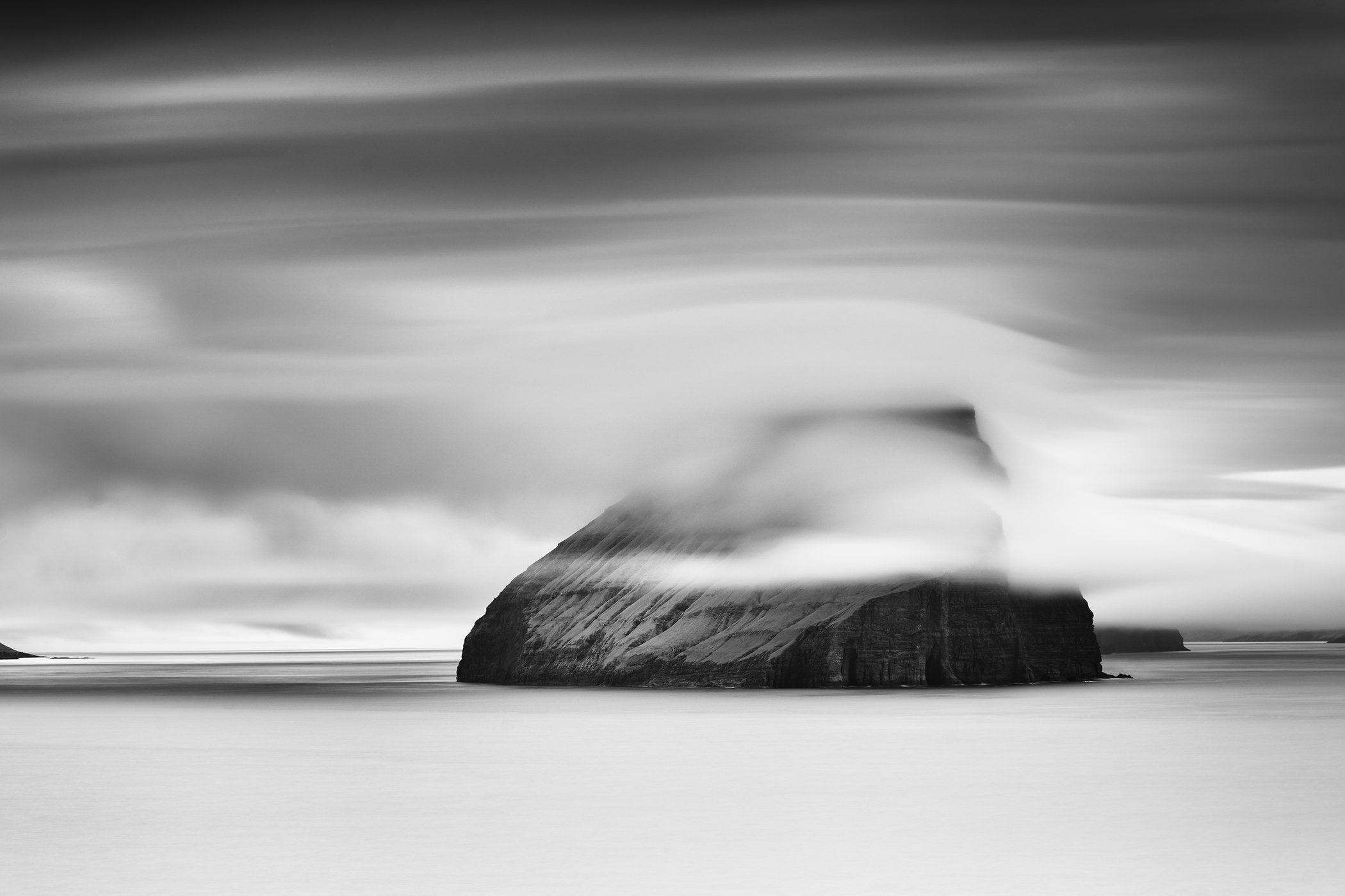 Black and white long-exposure photo of a sea stack in the Faroe Islands under low drifting clouds