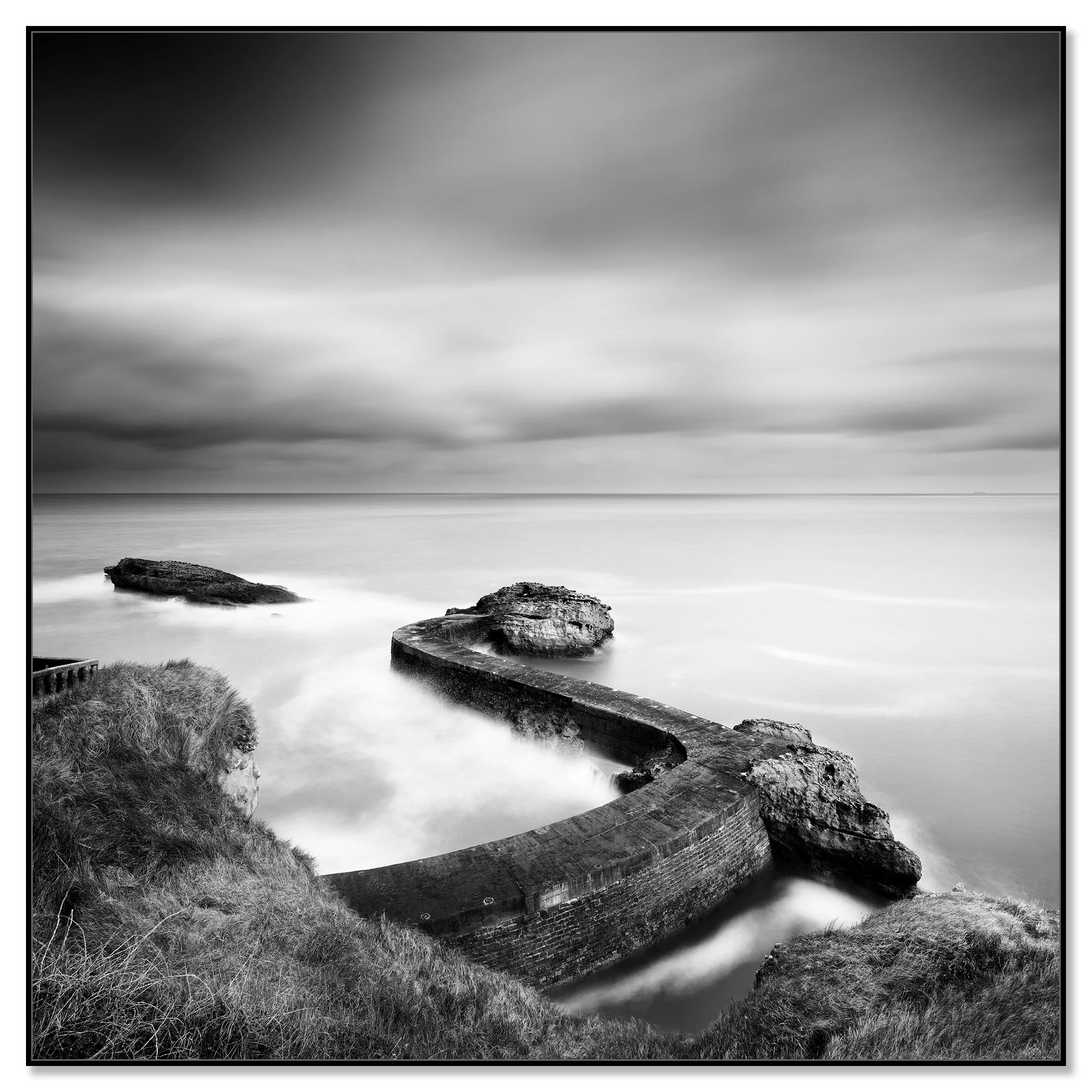 Black and white long-exposure seascape with a curved breakwater pier and rocks on a cloudy coast – framed ArtBox black