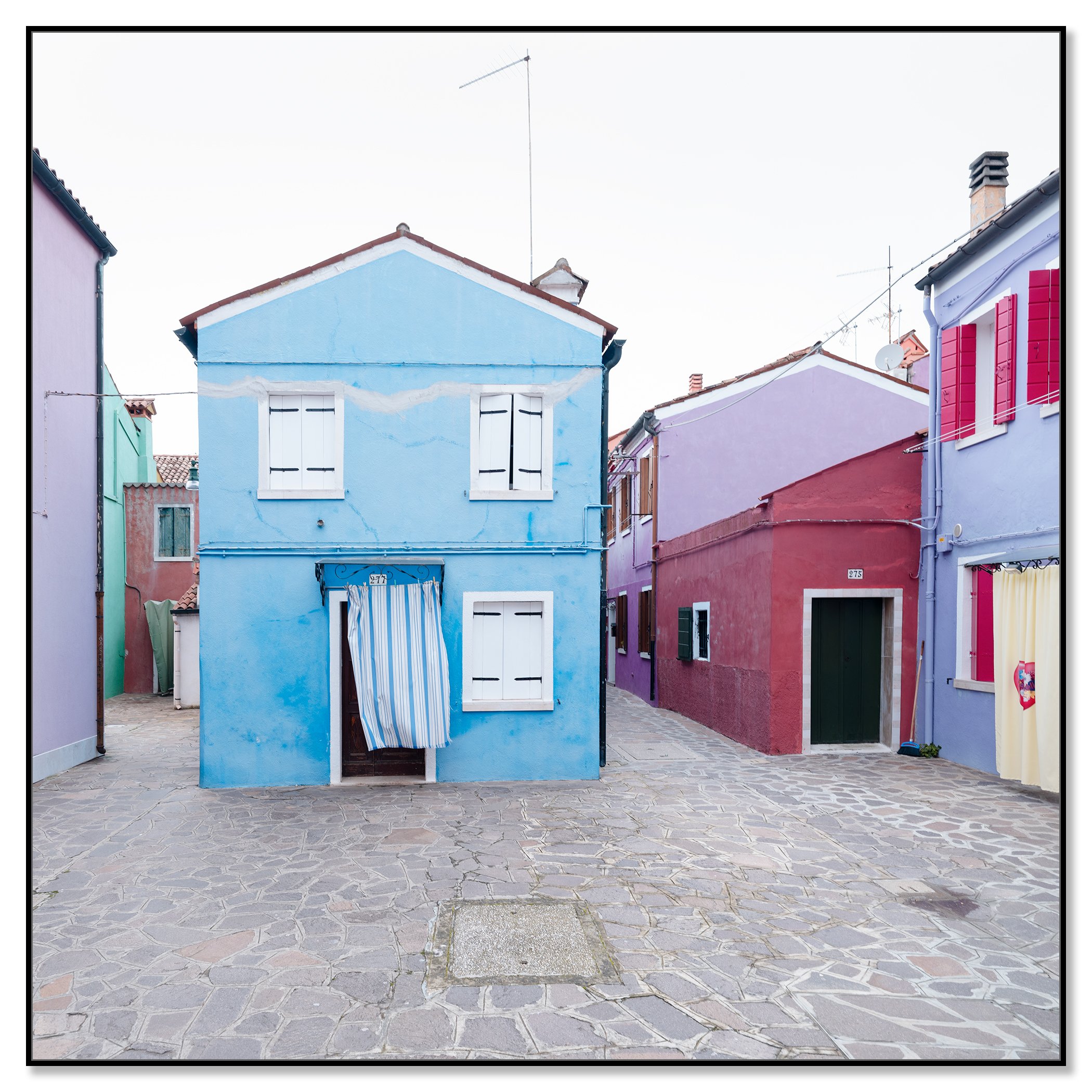 © 2025 Gerald Berghammer photo - Colorful houses in pastel shades on a cobblestone street, with colorful windows and a striped curtain hanging in front of one house. Chromaluxe framed black