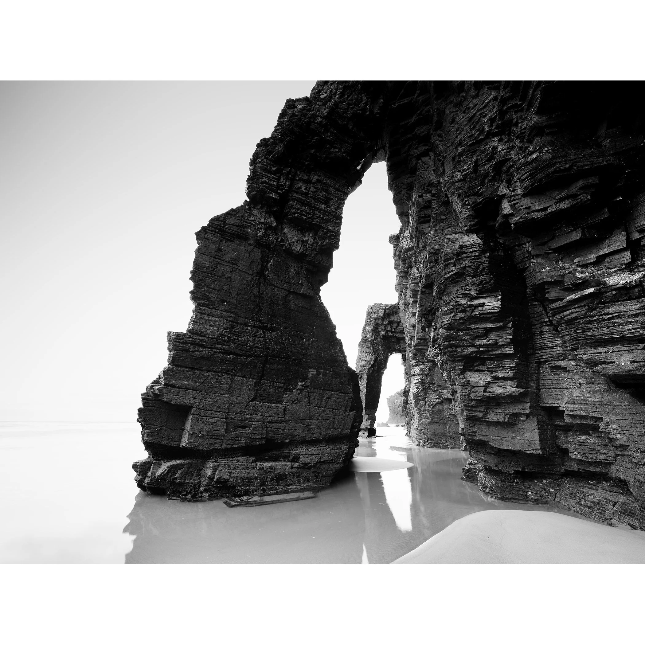Gerald Berghammer - Black and white beach seascape photography. Large natural rock arch formations on a beach with water reflecting the rocks.