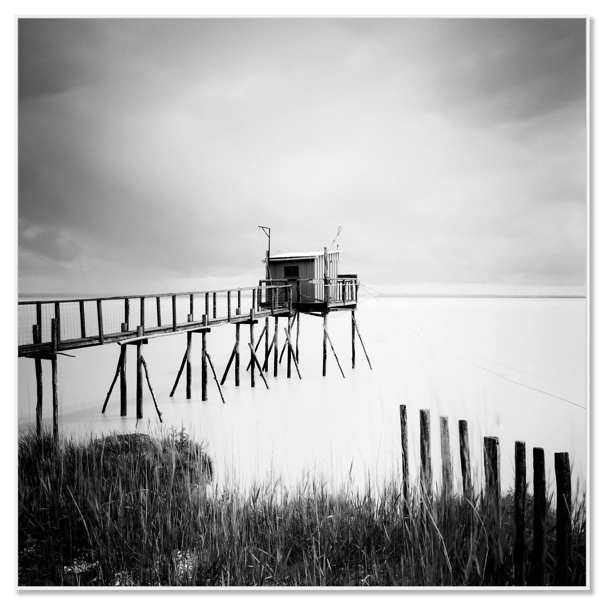 Gerald Berghammer - Black and white seascape photography. Small wooden hut on stilts connected by a long walkway over water with grass and a cloudy sky. Chromaluxe framed white