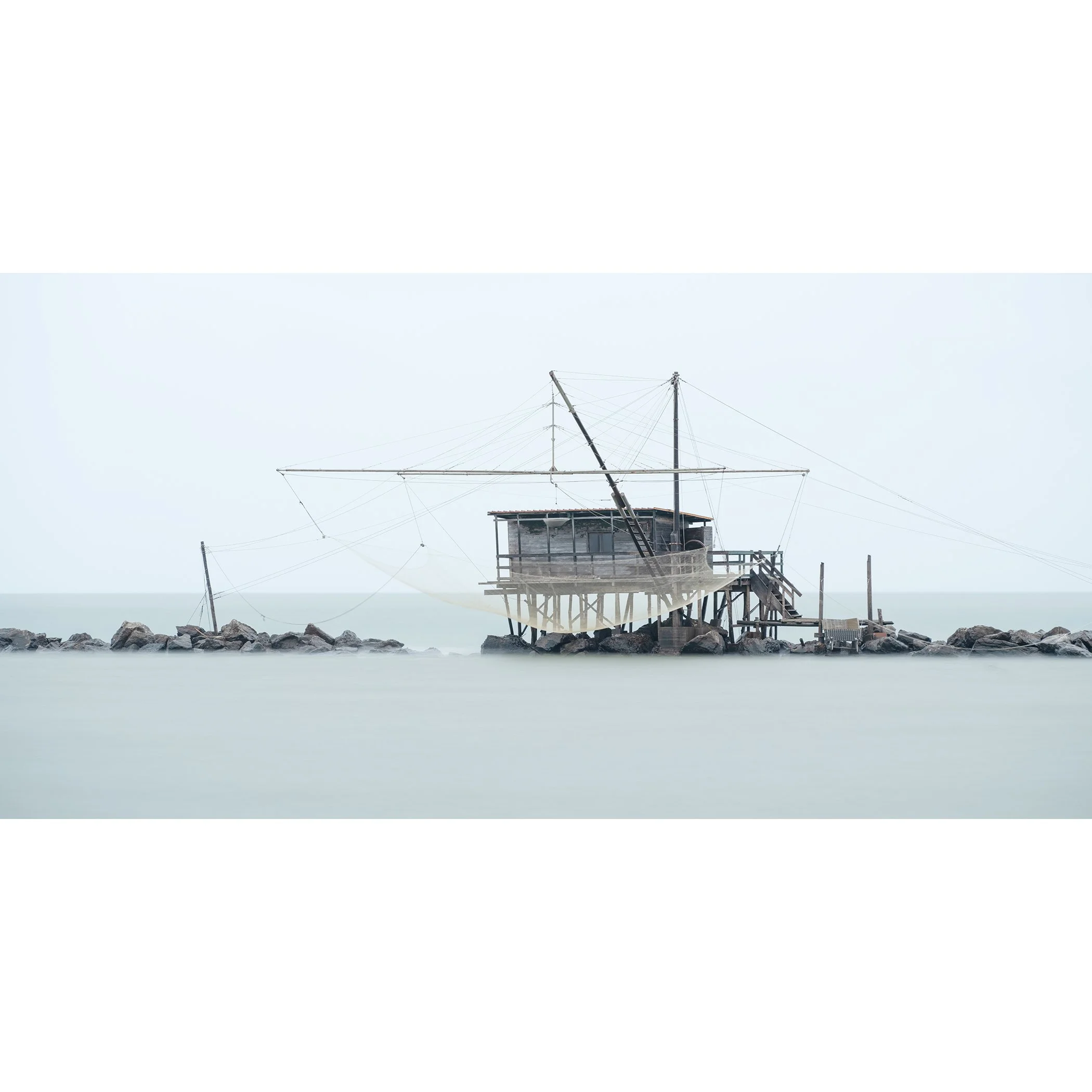 Gerald Berghammer - Color seascape photography. A rustic house on stilts with a crane and fishing nets, situated on rocks in the water, under a cloudy sky.