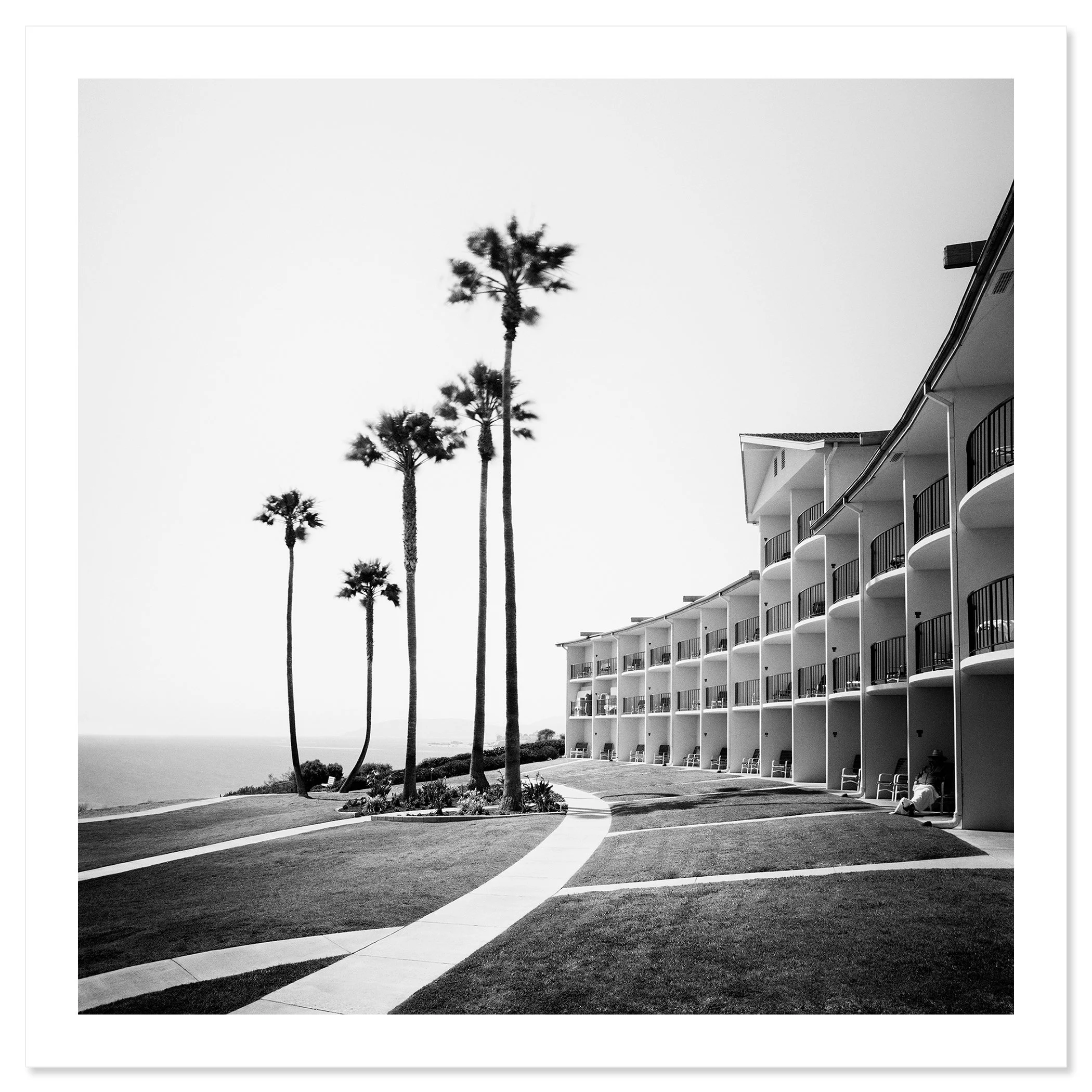 Gerald Berghammer - Black and white landscape photography. Seaside hotel with multiple balconies, a curved walkway, and tall palm trees near the ocean. Fine art print only