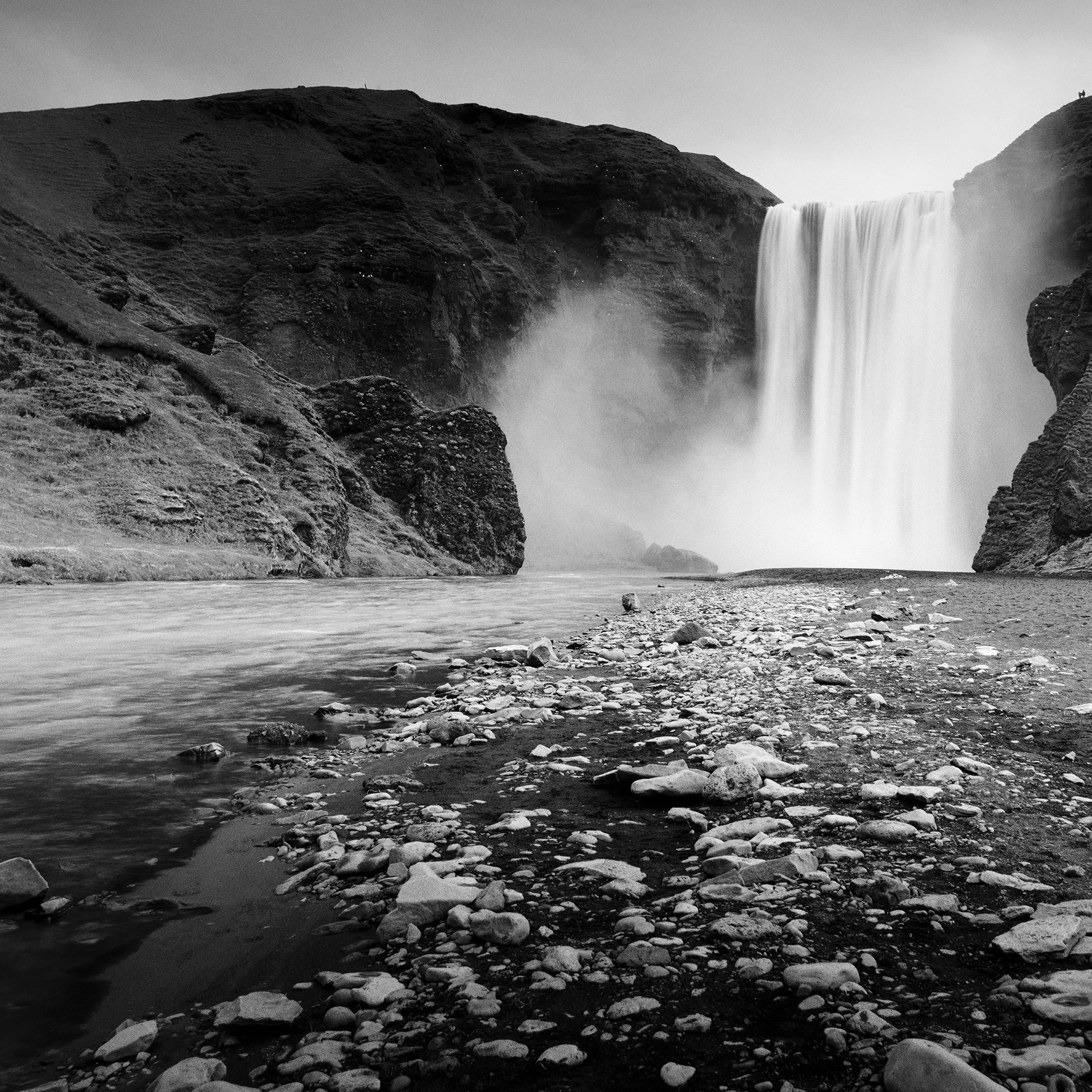 Gerald Berghammer - Black and white long exposure waterscape photography. A large waterfall flowing between rugged cliffs into a rocky river. Print detail 1