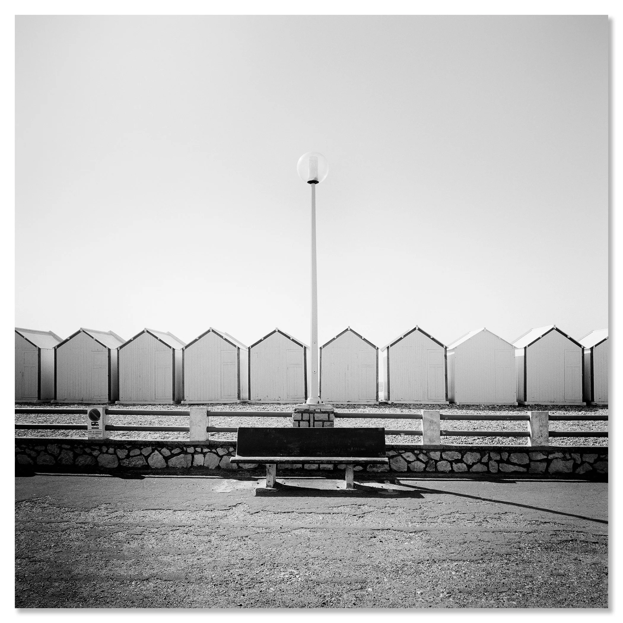 Empty bench on promenade facing beach huts, centred streetlamp; minimalist black-and-white coastal photo – dibond frameless