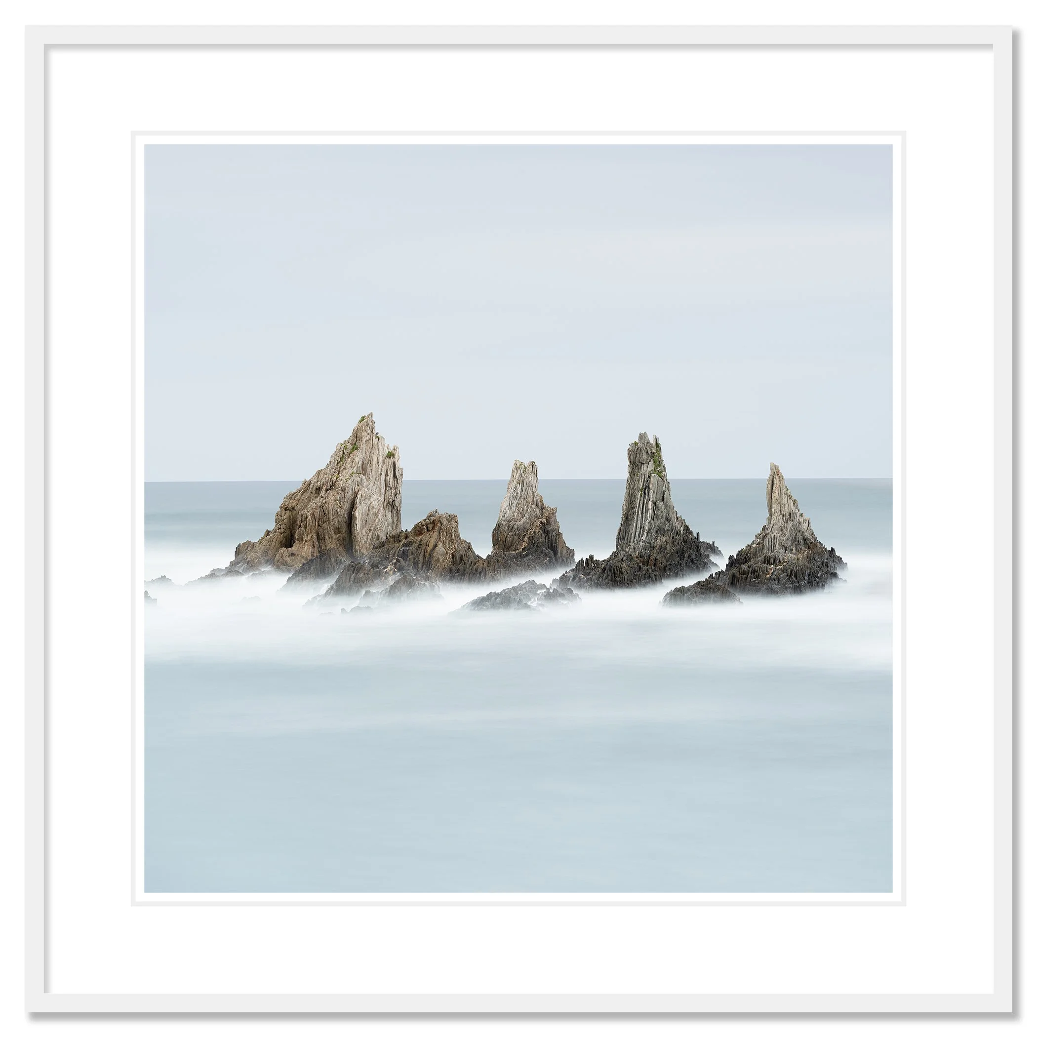 Gerald Berghammer - Color long exposure fine art photography. A seascape with five tall, jagged rocks emerging from the water and a hazy, blue sky. Classic framed white