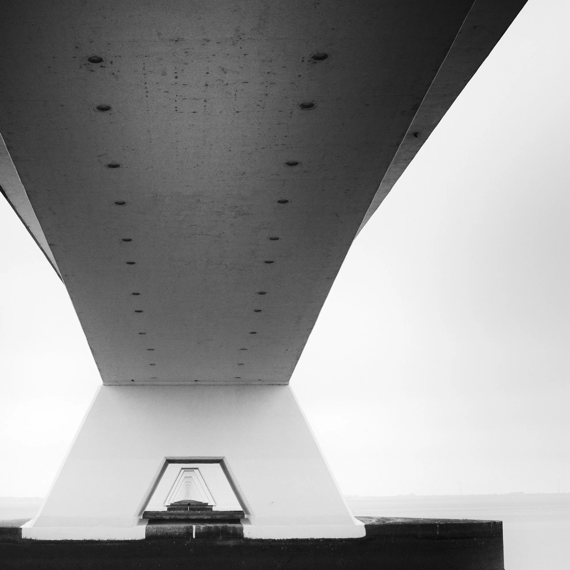 Gerald Berghammer - Black and white cityscape photo. Underneath view of a bridge with large support pillars, showing the underside and the water below. Print detail 2