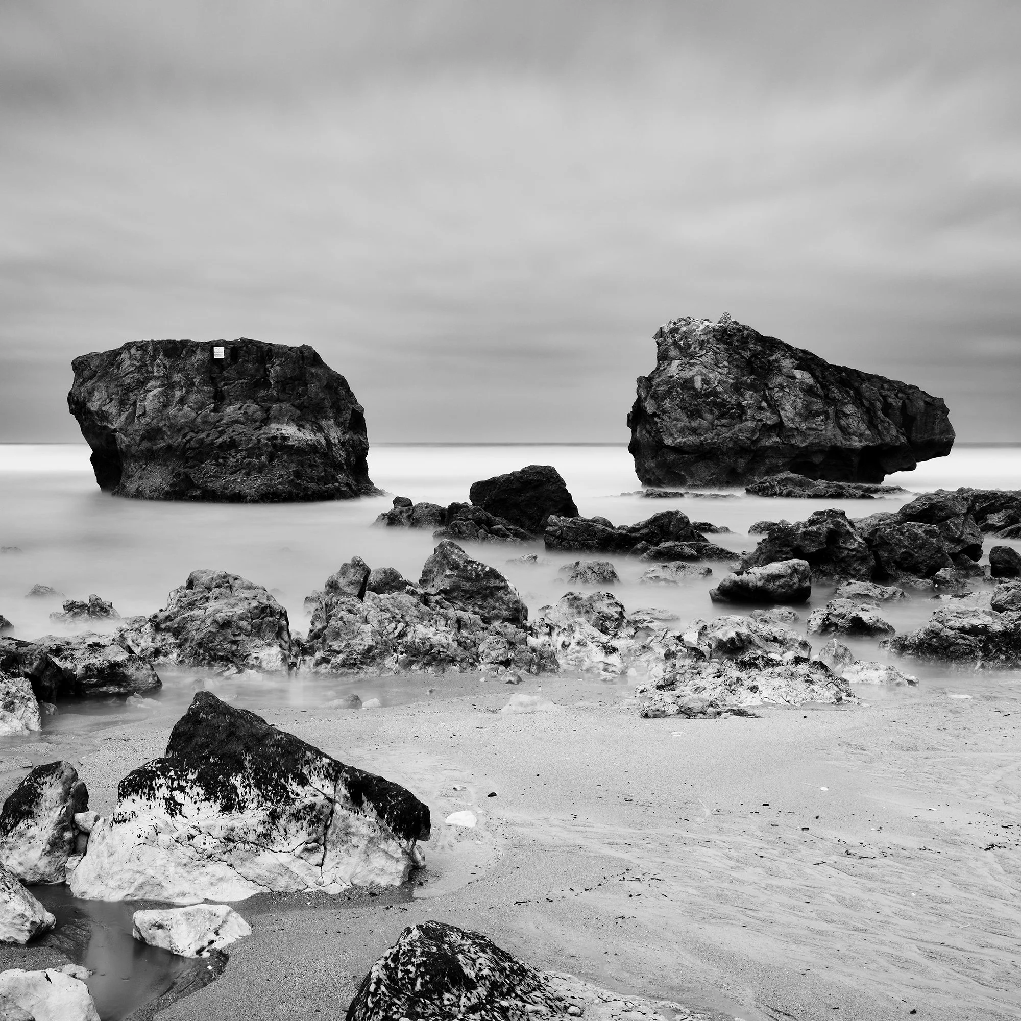 Gerald Berghammer - Black and white landscape photo. Rocky beach with two large rocks protruding from the water, cloudy sky, and smooth ocean surface. Print detail 3