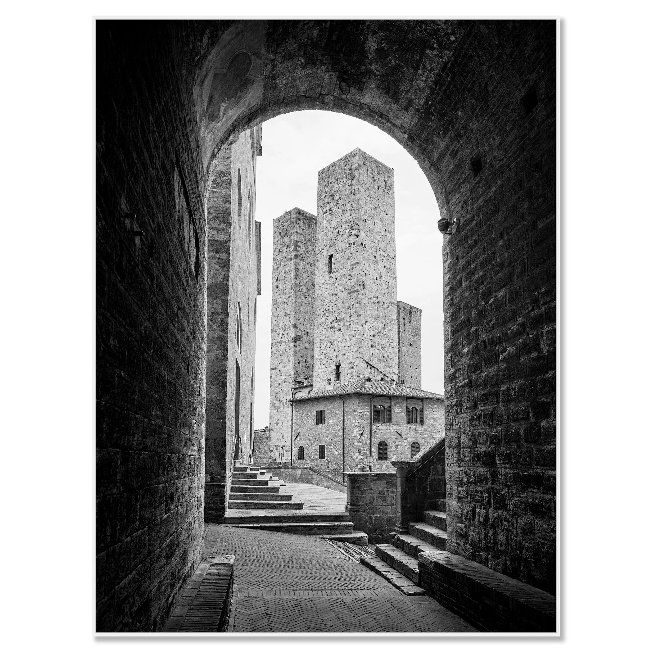Black-and-white photograph of a medieval stone tower and buildings in San Gimignano, viewed through an arched stone gate – framed ArtBox white