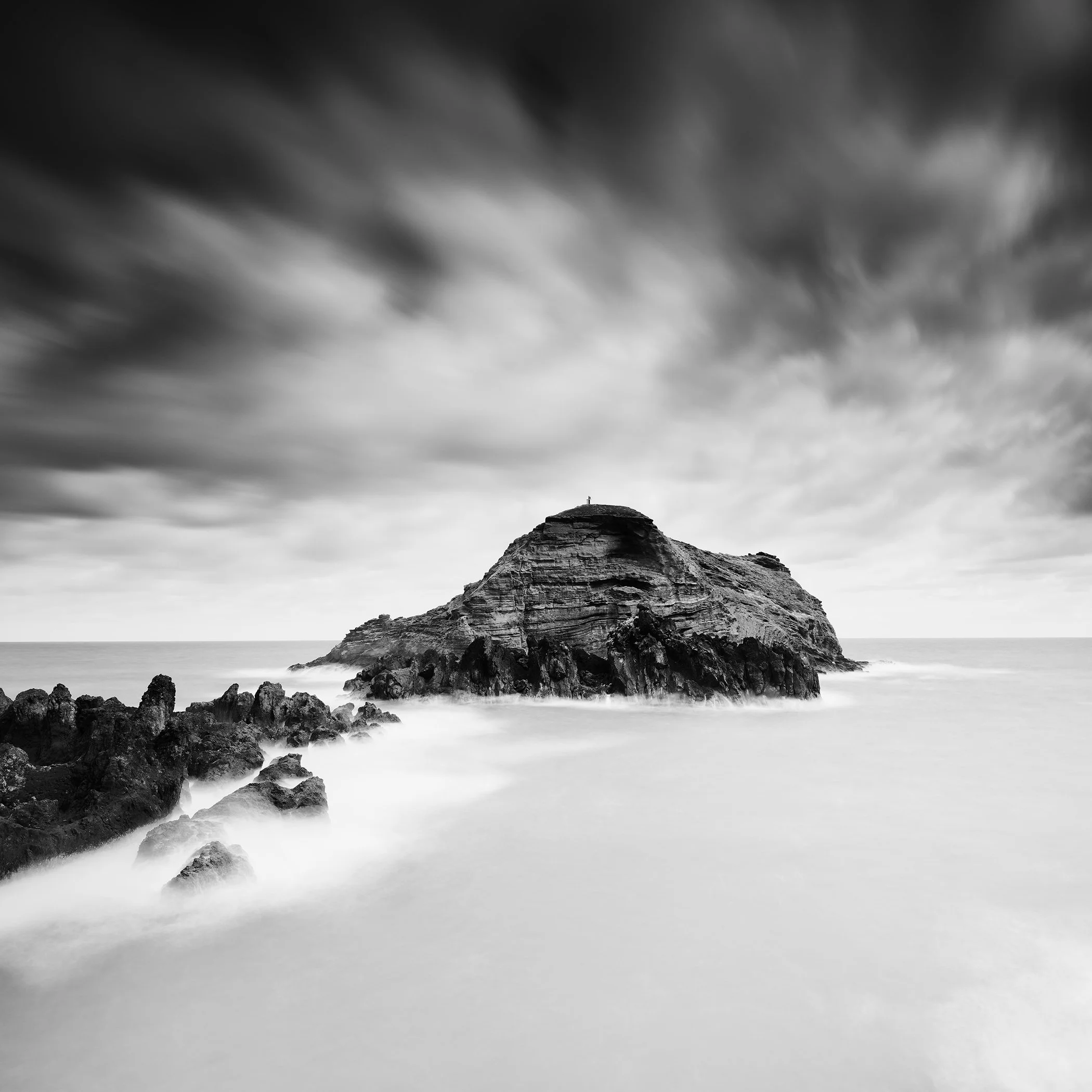 Black-and-white long-exposure seascape of a rocky island beneath dramatic clouds.