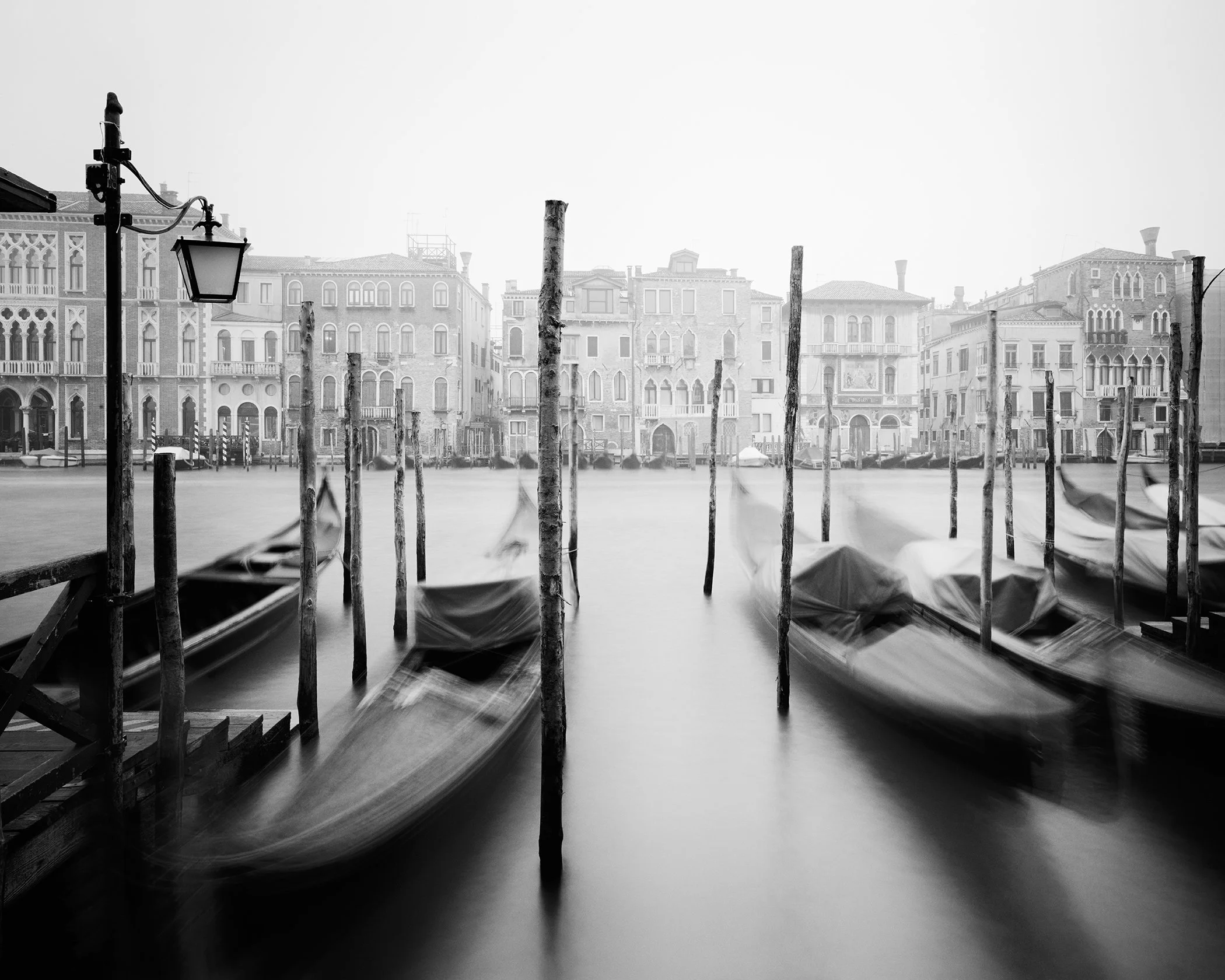Black-and-white long-exposure photograph of gondolas and wooden mooring poles on Venice’s misty Canal Grande. Photo by Gerald Berghammer.