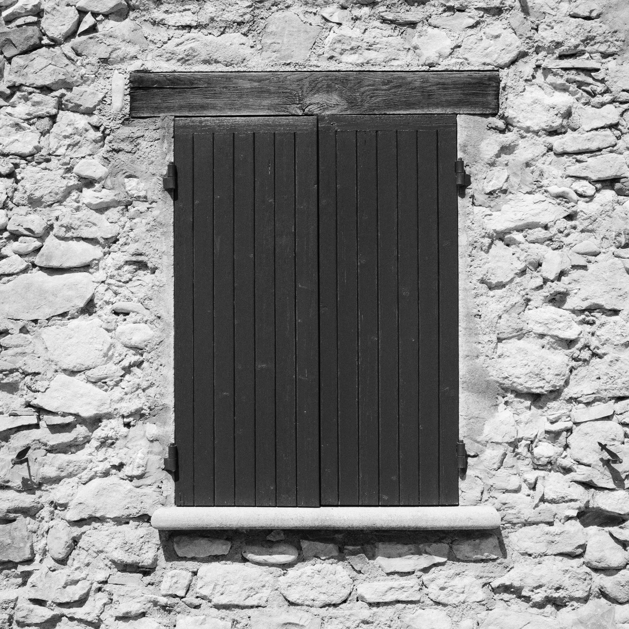 Gerald Berghammer - Black & white photography. Stone wall with a closed wooden window shutter, flanked by climbing vines on both sides, and a small bench. Print detail 3