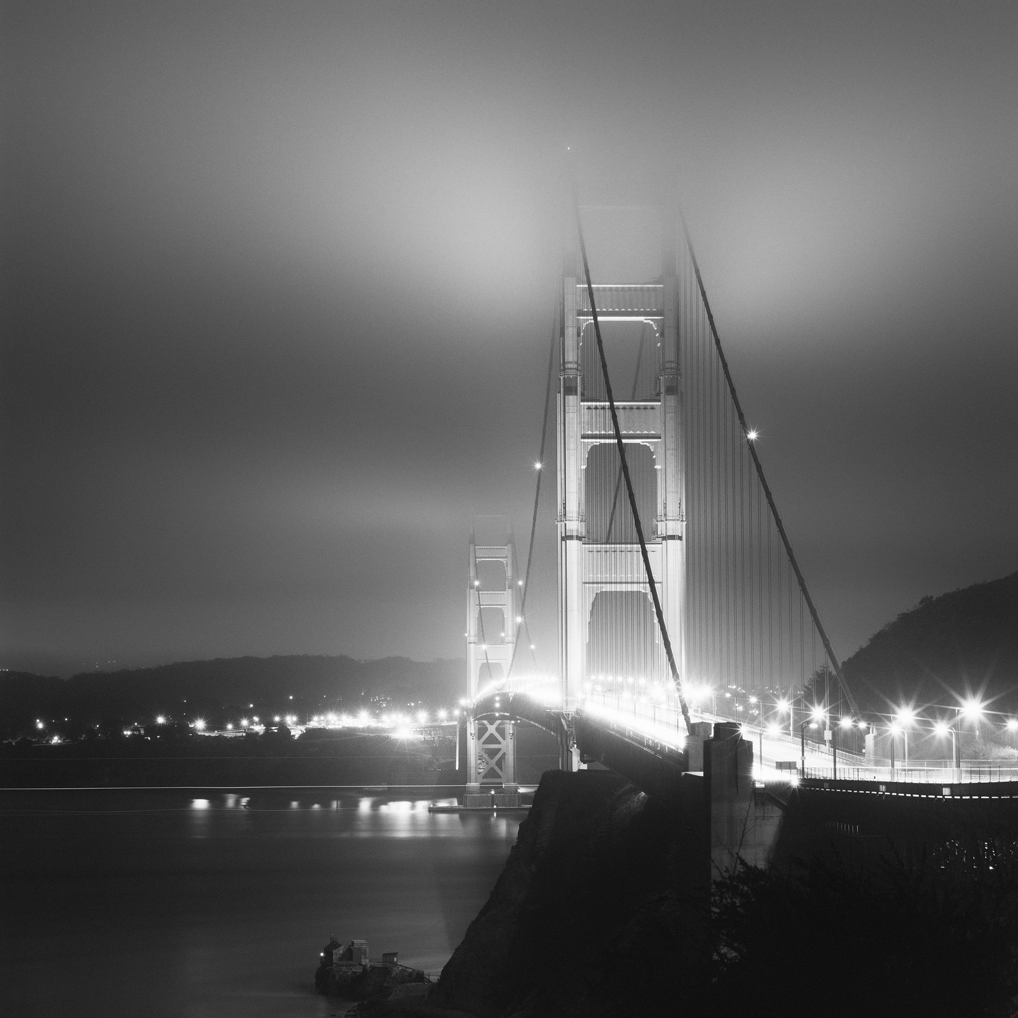 Black-and-white night photograph of the Golden Gate Bridge illuminated through fog above San Francisco Bay by Gerald Berghammer.