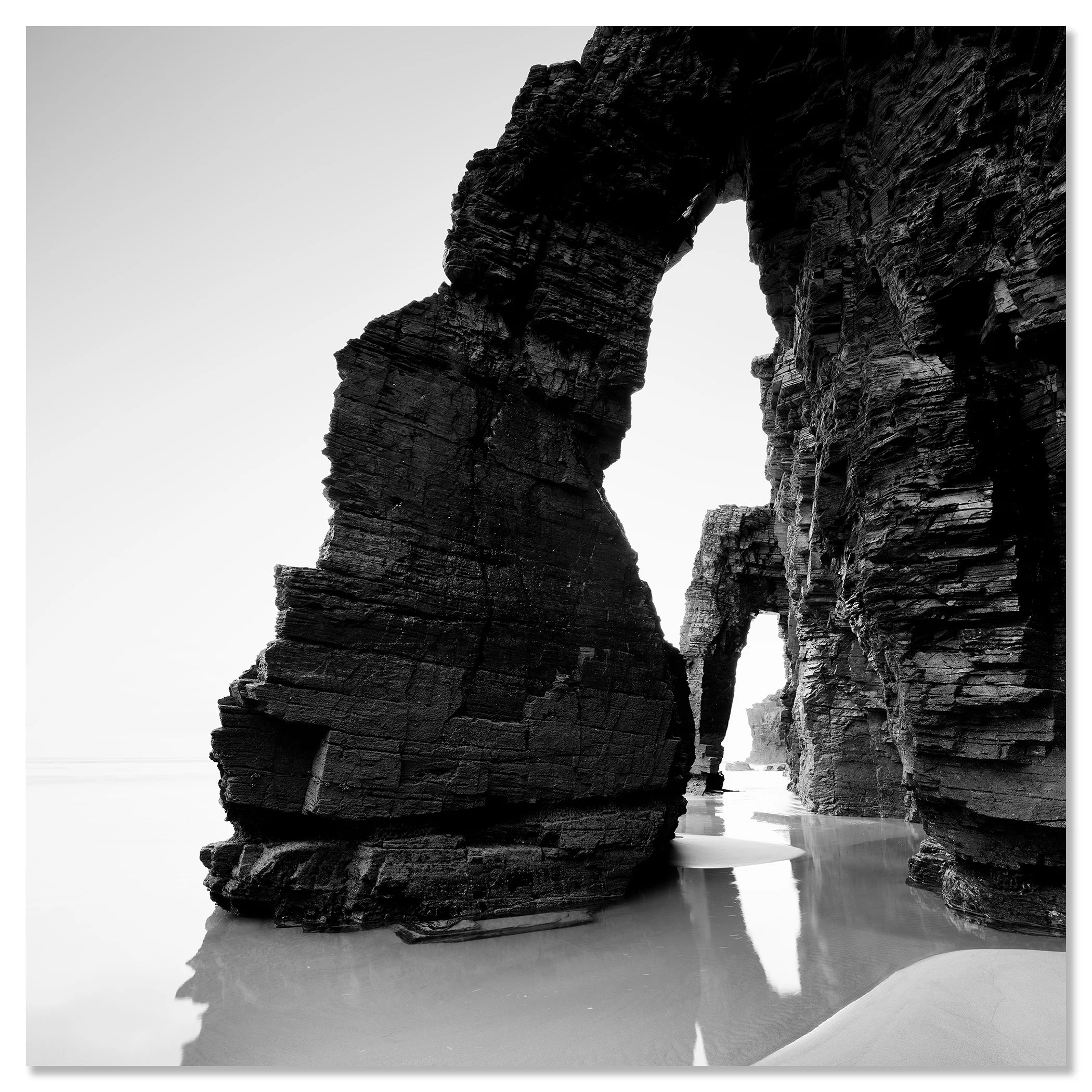 Black and white photo of dramatic sea arches and layered rock cliffs on a quiet sandy beach – dibond frameless