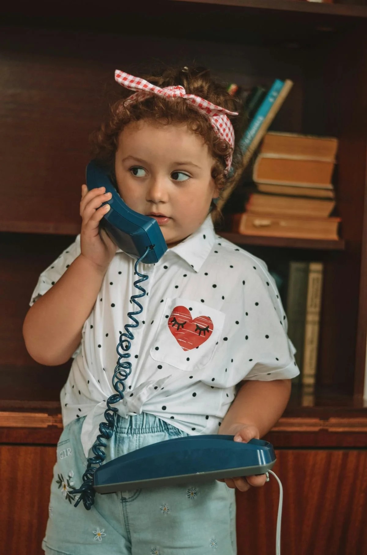 A young girl is standing in front of a bookshelf holding a blue phone to her ear looking sideways