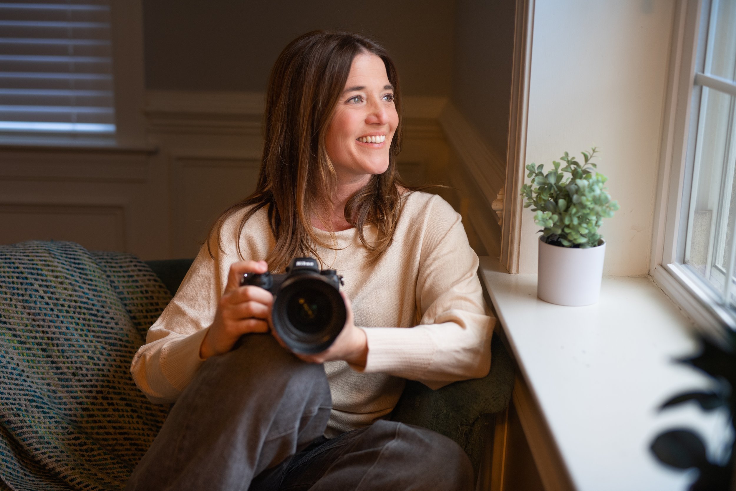 A woman sitting by a window holding a camera, smiling, with a potted plant on the windowsill.