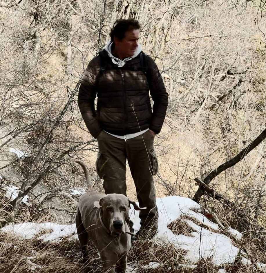 Un uomo con una maglietta a maniche lunghe, una giacca e pantaloni cargo cammina in una foresta innevata con un cane di taglia grande, in un ambiente naturale con alberi spogli.