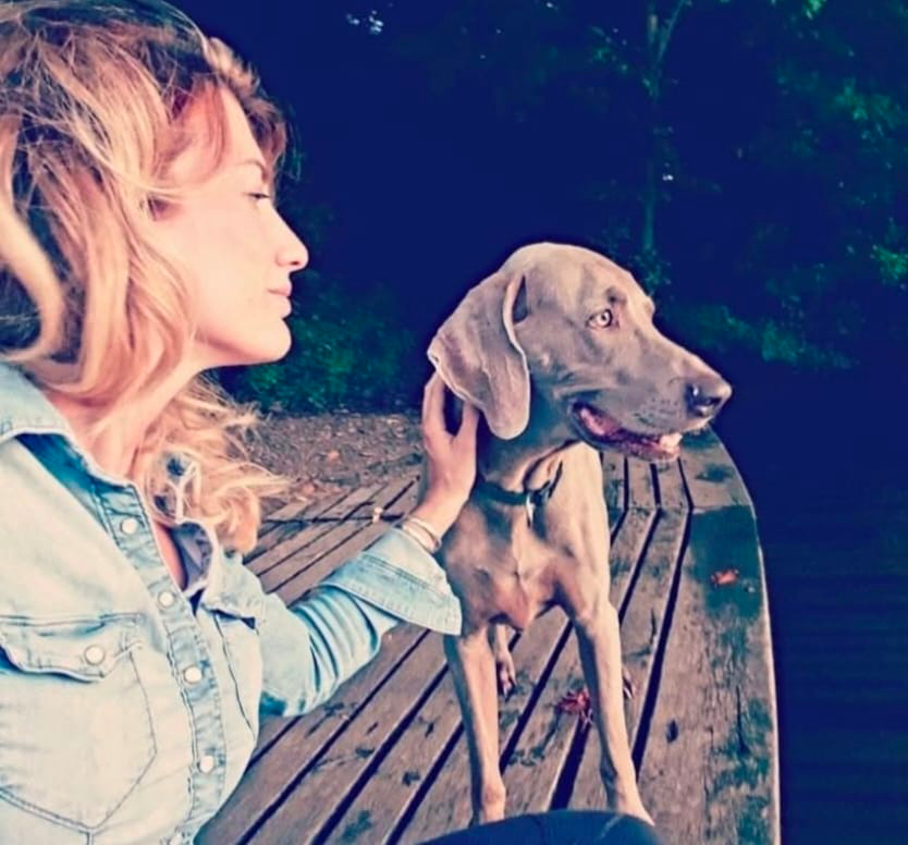 Donna con capelli ricci biondi e giacca di jeans accarezza un levriero grigio su un ponte di legno circondato da natura.