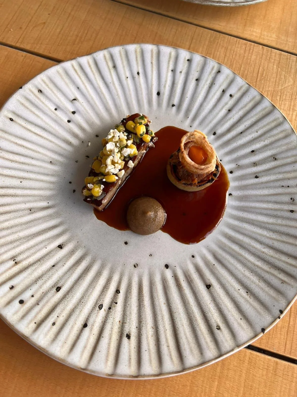 A modern plated appetizer with a rectangular piece topped with popcorn, a small round item, and a cylindrical roll with an orange filling, all on a decorative white plate with black specks.