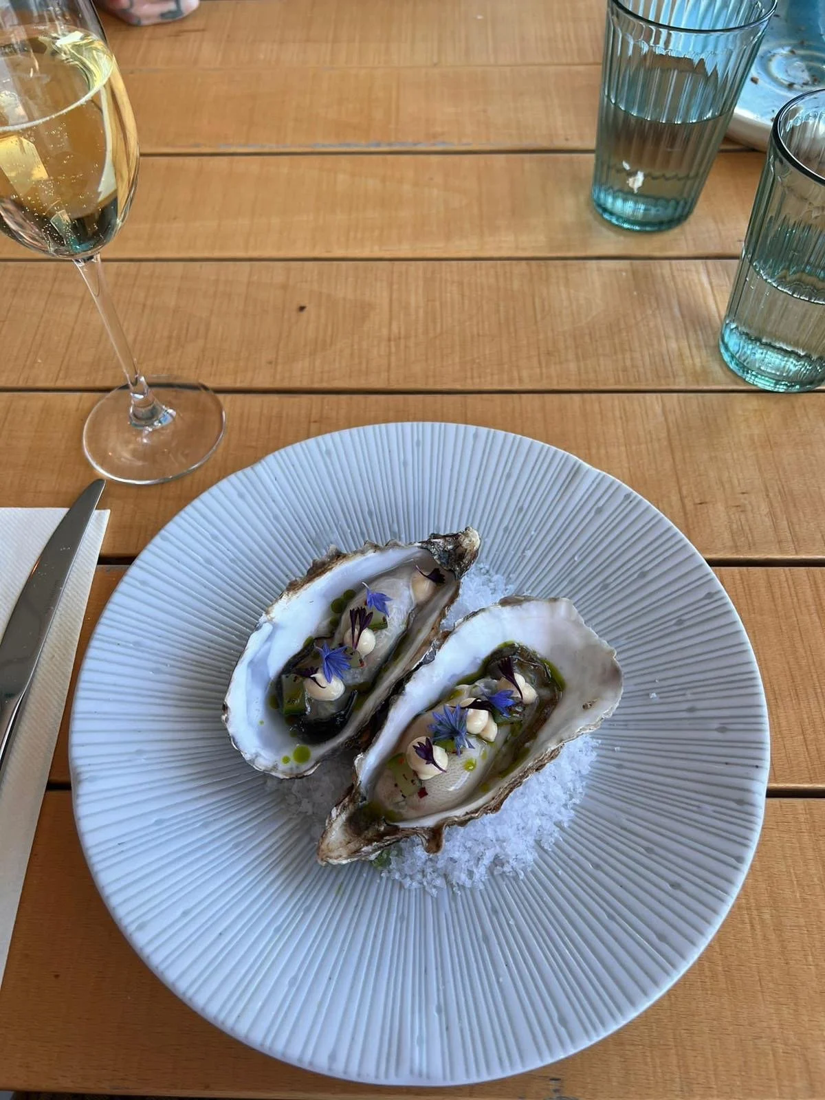 Two oysters on a white plate garnished with edible flowers, herbs, and sauces, placed on ice, on a wooden table with a glass of white wine and several glasses of water nearby.
