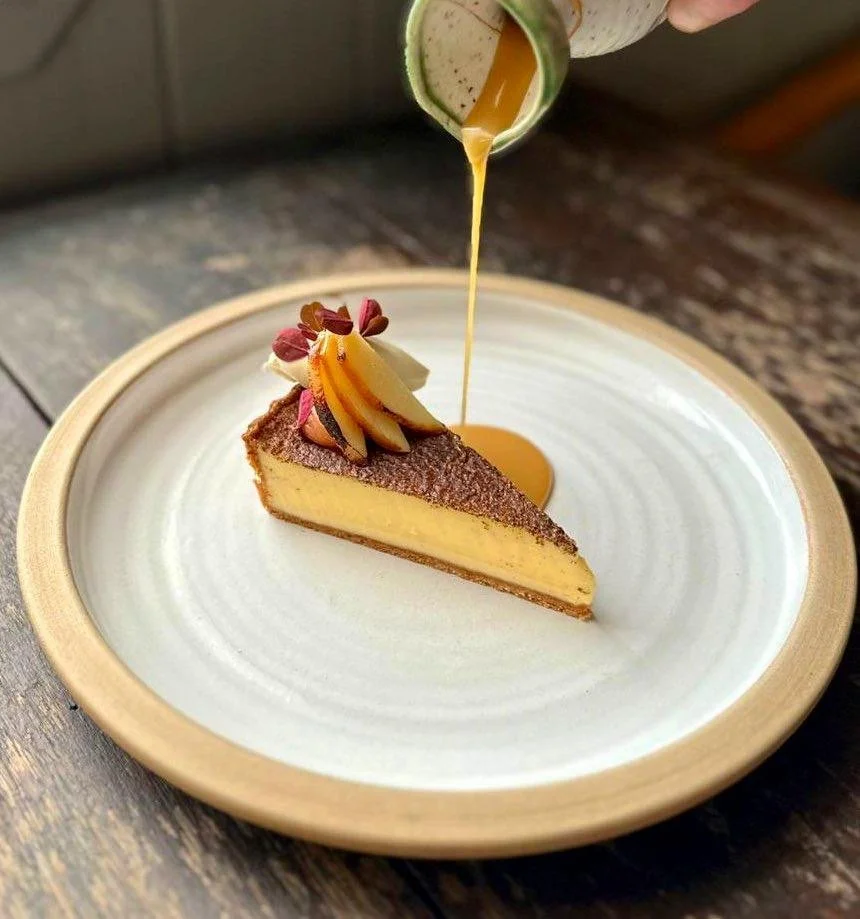 A slice of cheesecake on a white plate with a decorative crust, topped with whipped cream, apple slices, and edible flower petals. Caramel sauce is being poured over the cake from a small container.
