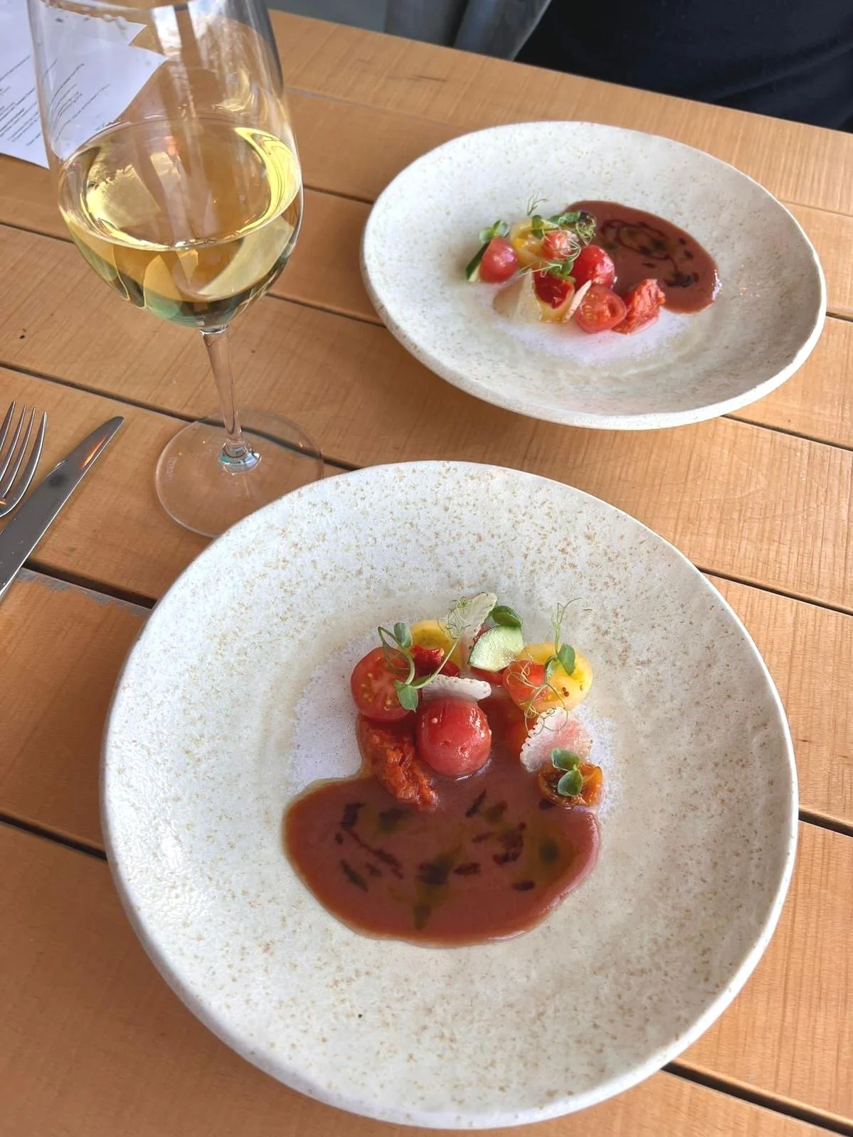 Two white plates with gourmet salads and a glass of white wine on a wooden table.