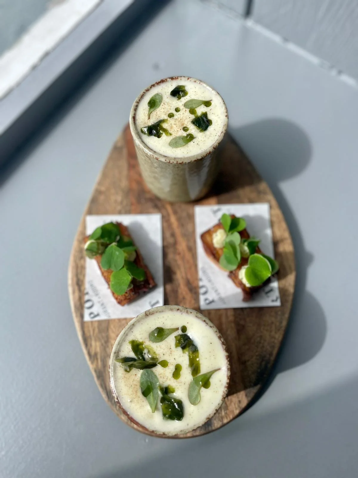 Two tall glasses of a creamy beverage with green herbs or toppings, placed on a round wooden serving board with two small rectangular pieces of toast garnished with greens.