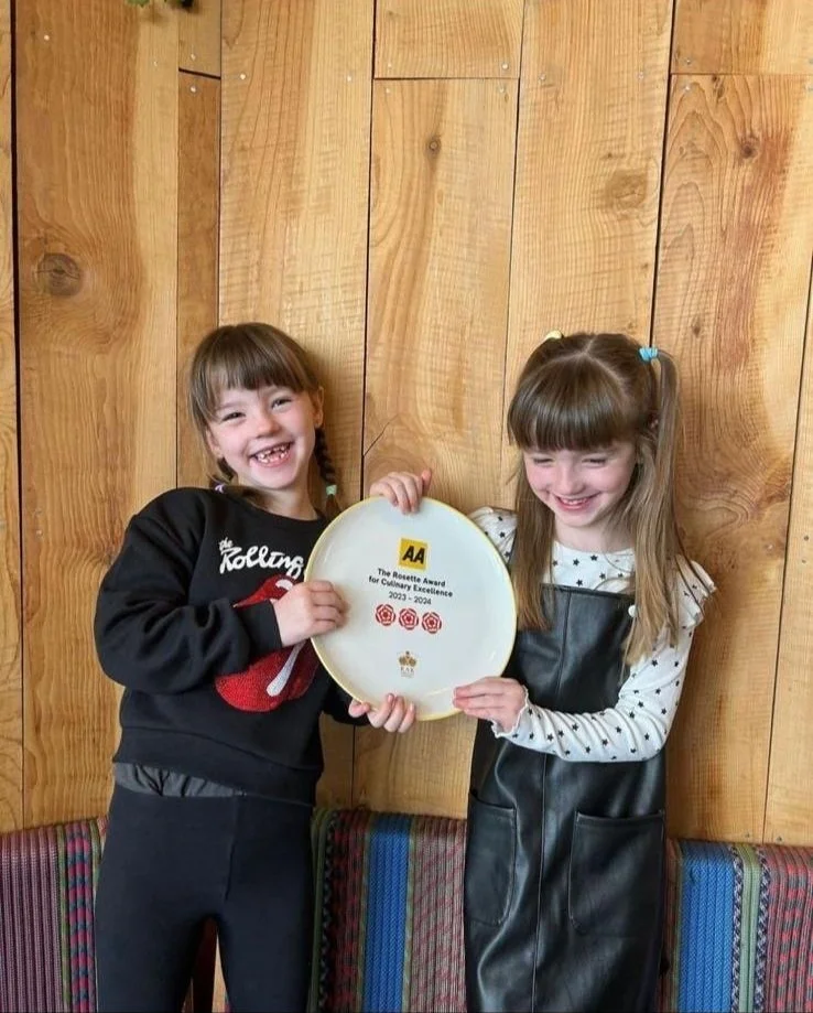 Two smiling young girls holding a white plate with a logo and text, standing in front of a wooden wall, with colorful upholstered seating behind them.