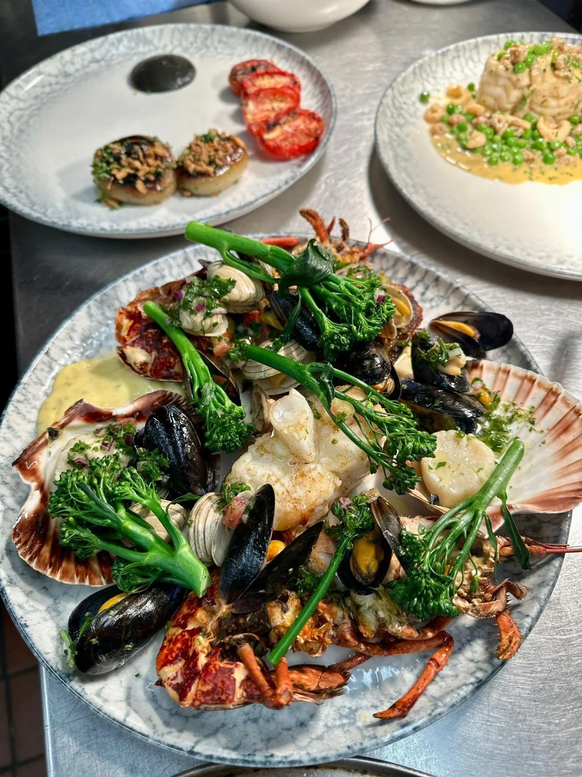 Seafood platter with lobster, mussels, scallops, and green herbs with three other dishes in the background, on a table.