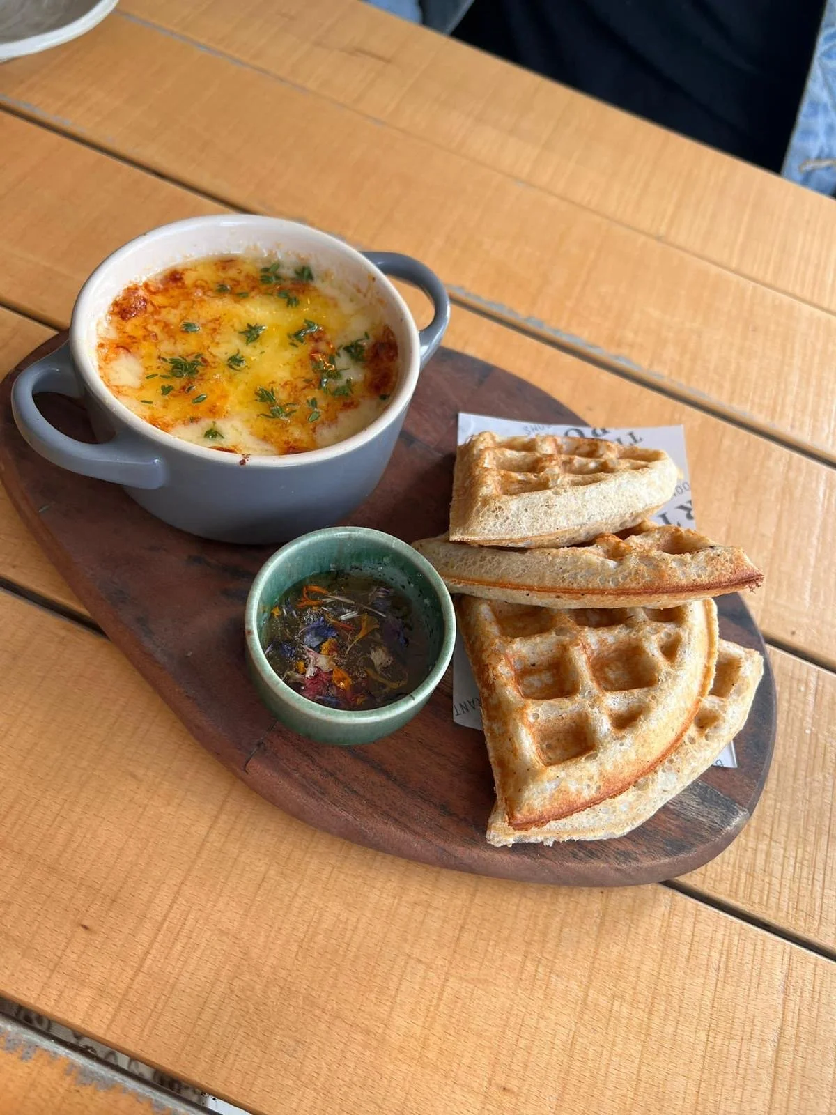 A serving of baked eggs with cheese and herbs, three pieces of waffle, and a cup of hot sauce on a wooden platter.
