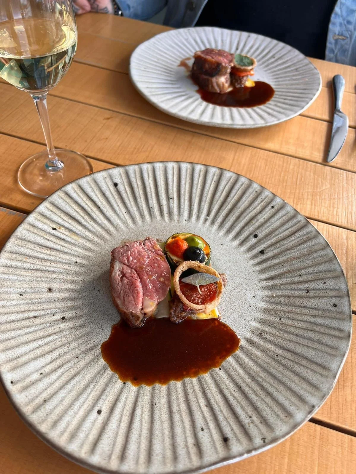 A gourmet plate featuring a piece of pink, medium-rare steak, a dollop of dark sauce, a small omelet with cherry tomato, black olive, and green garnish, and a crispy onion ring, served on a textured ceramic plate.