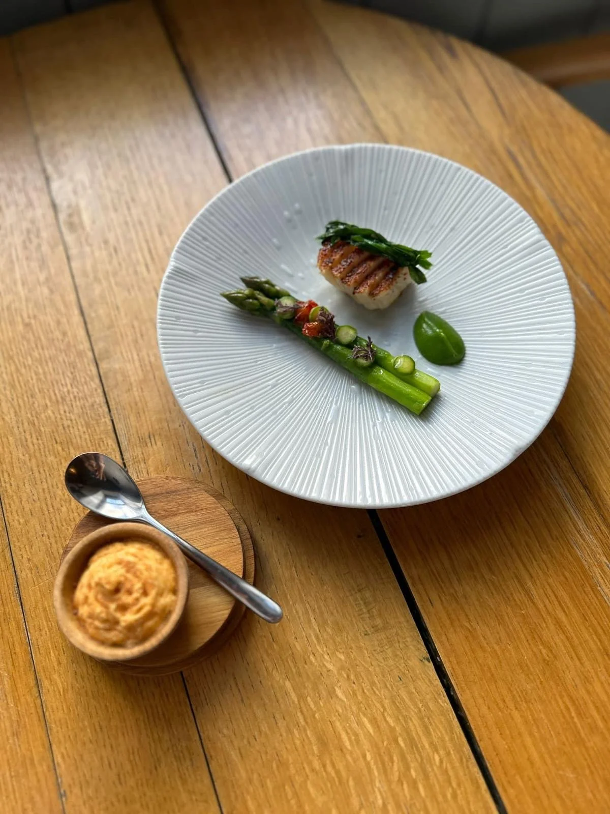 A white textured plate with a piece of grilled fish topped with greens, a stalk of asparagus with spices, and a green sauce, on a wooden table. A small bowl with a spoon and some food is beside the plate.