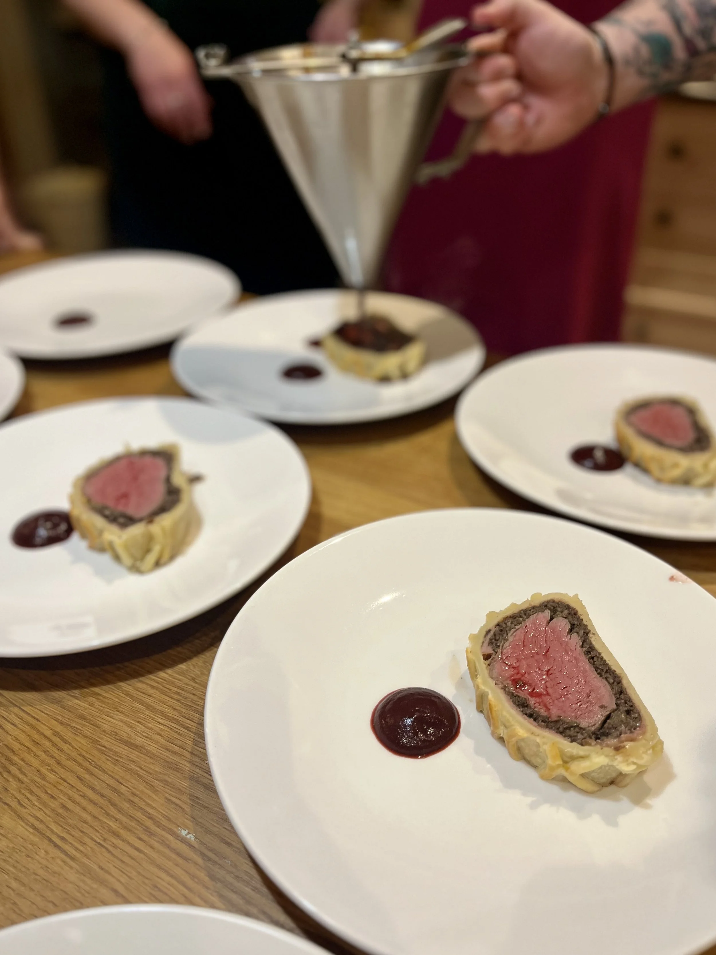 Slices of Beef Wellington on white plates with a dollop of red sauce, arranged on a wooden table. A person with tattoos is pouring sauce from a metal gravy boat in the background.