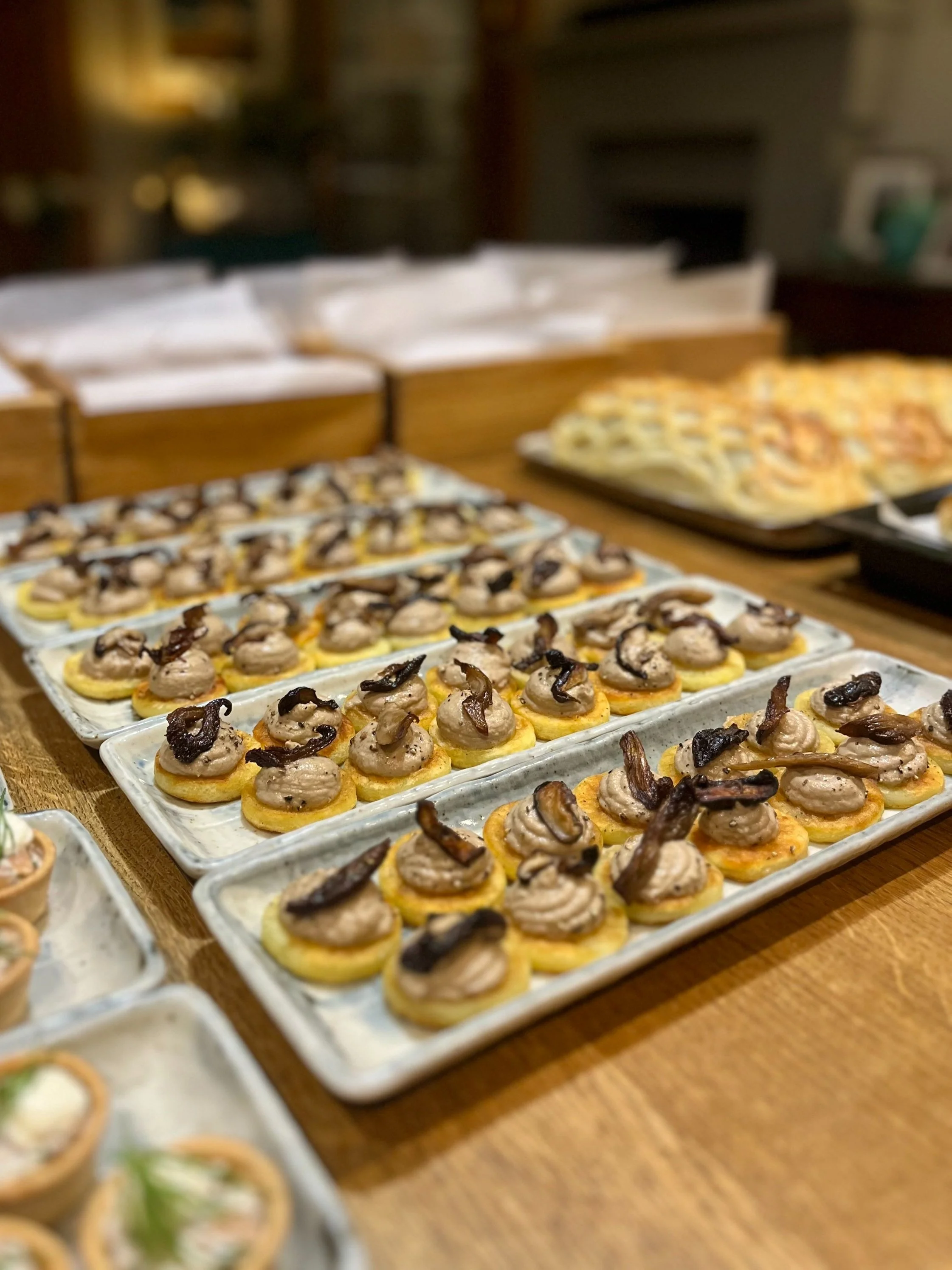 Tray of bite-sized appetizers topped with a dollop of spread and crispy bacon pieces on a wooden table at a party or gathering.