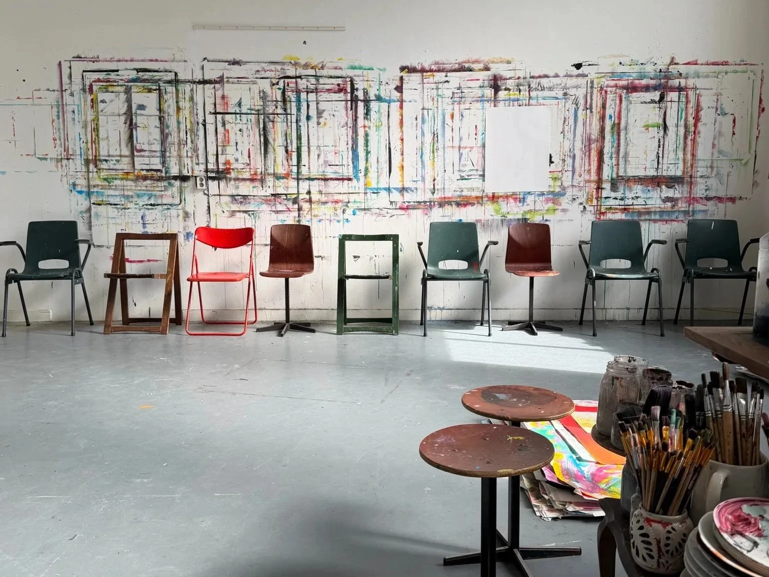Een kunststudio met acht stoelen en een muur met abstracte tekeningen, en een tafel met verfpotten, brushes en papier. Een ruimte om te experimenteren, met verf op de muur.
