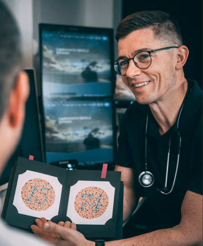 Sjømannslege med stetoskop rundt halsen undersøker sjømann med hjelp av en Ishihara fargesynstest og schnellen synstavle