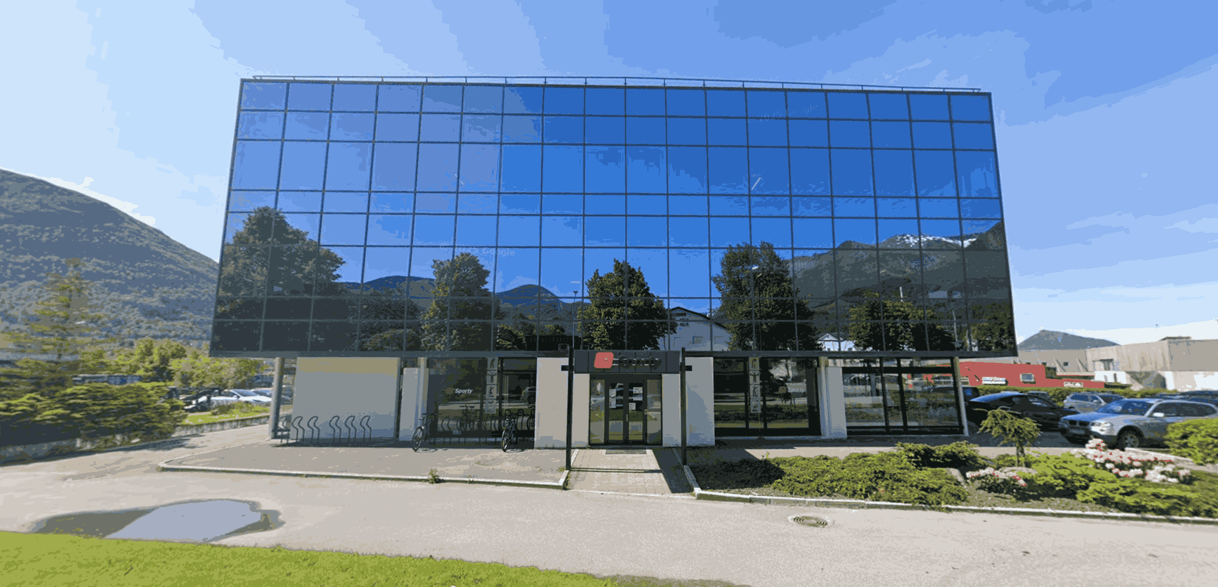 Modern glassbygg med refleksjonsvinduer, omgitt av parkering og grøntarealer med blomster og trær, omgitt av fjell og klar blå himmel.