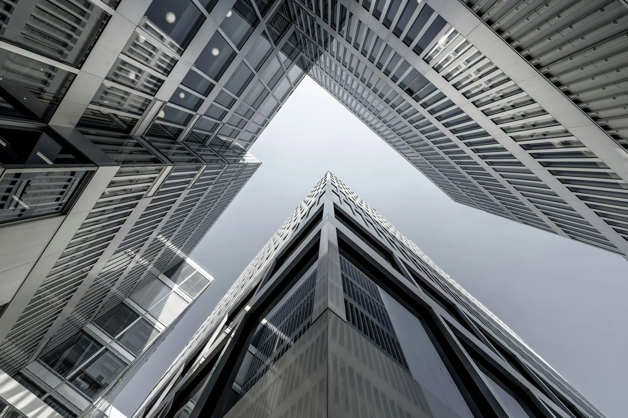 Looking up at tall modern skyscrapers in a city, with structural glass windows, metal frameworks, and a cloudy sky above.