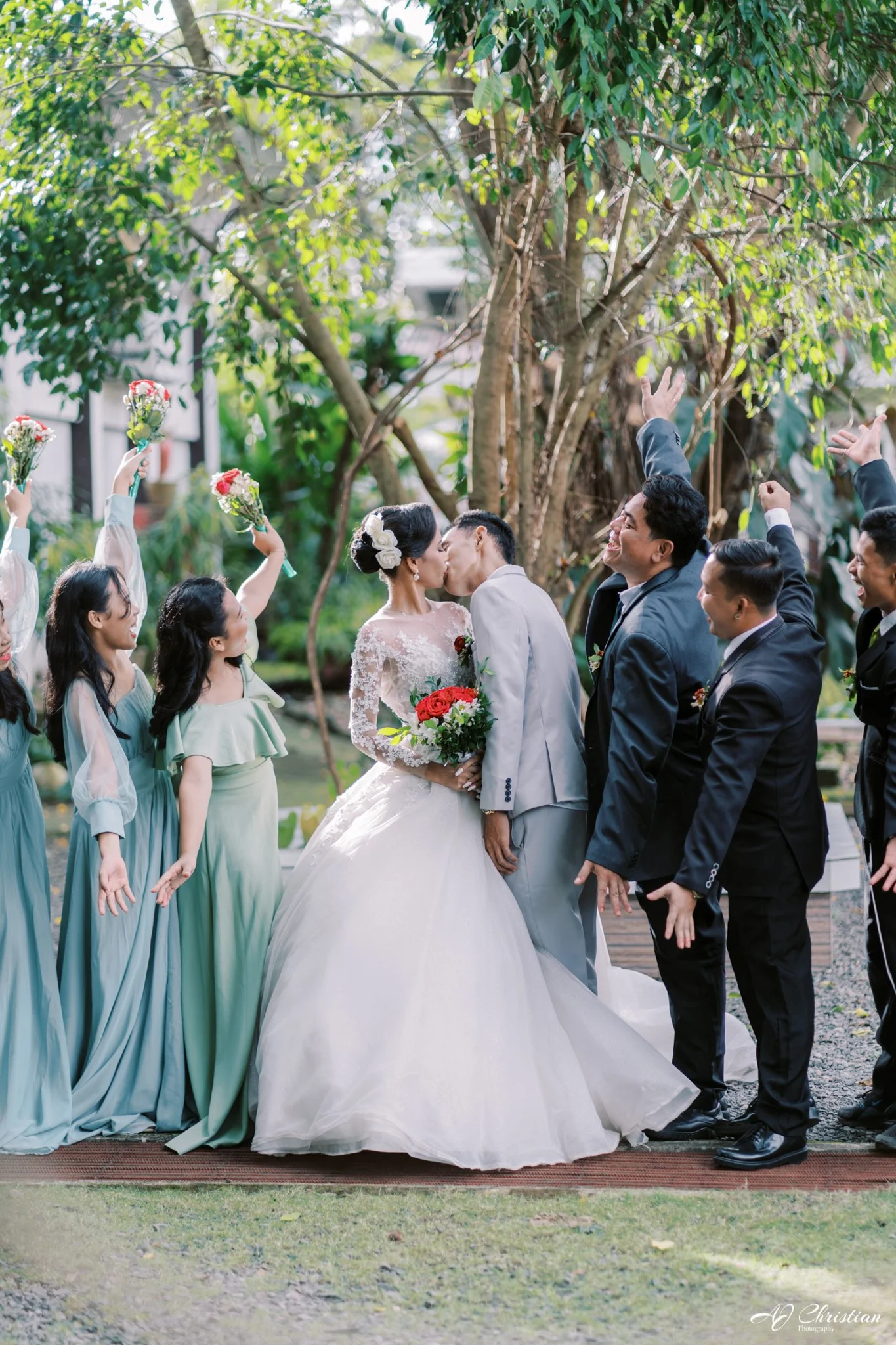 Bride and groom kissing surrounded by wedding party posing with raised hands outdoors.
