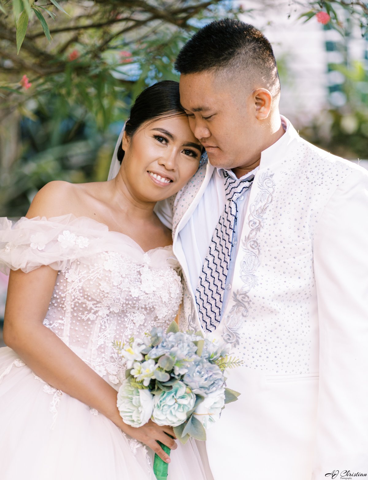 Bride and groom embracing in wedding attire holding a bouquet, surrounded by greenery.