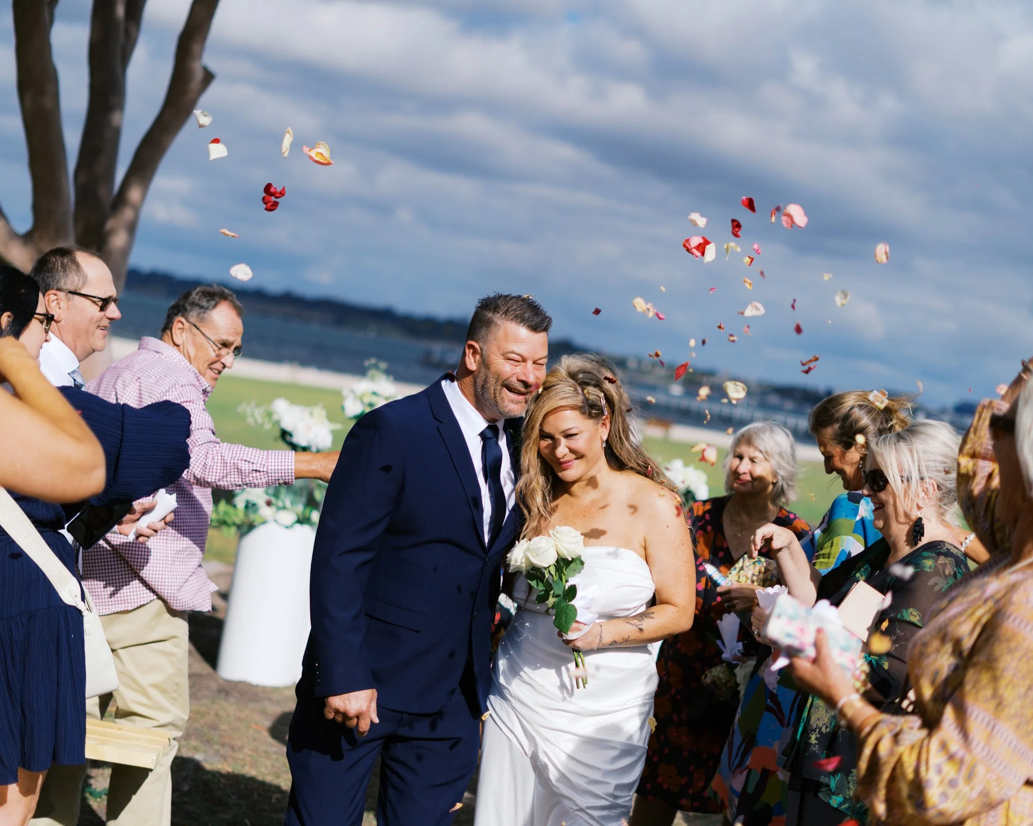 Joyful bride and groom showered with red and white petals by smiling wedding guests outdoors near a body of water under a partly cloudy sky.