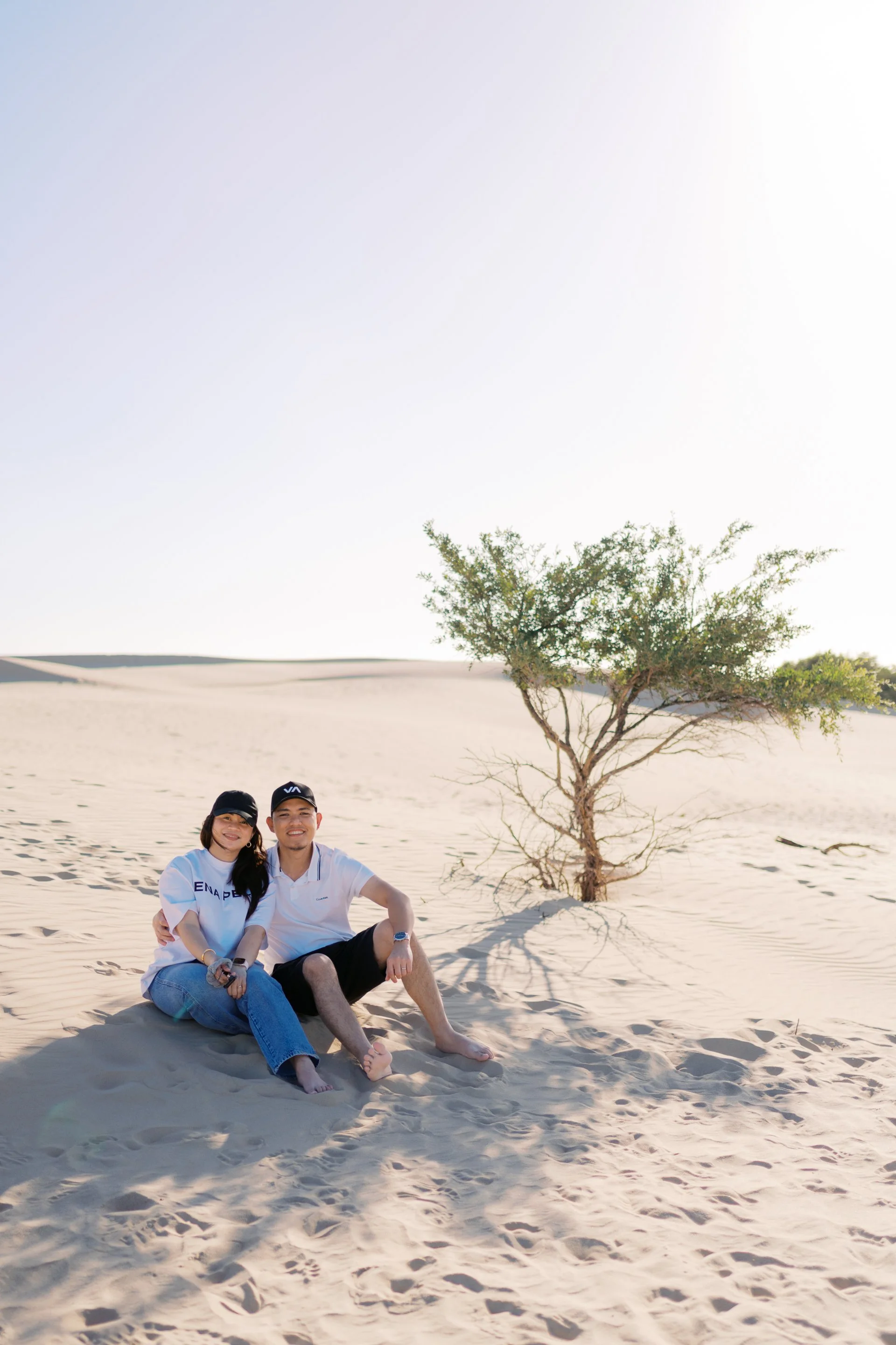 Two people sitting on desert sand next to a small tree in Big Drift, Wilsons Promontory, Victoria, Australia