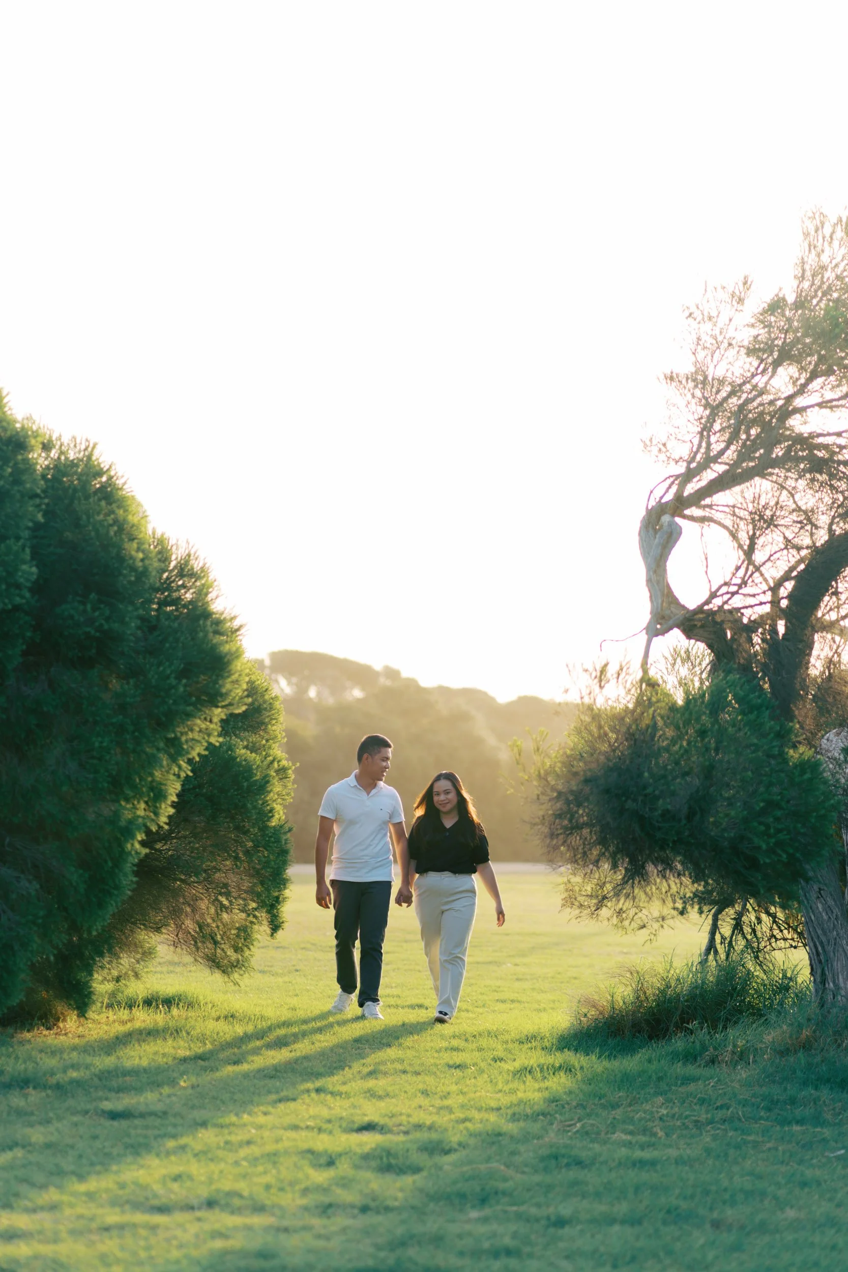 A couple walking hand in hand through a grassy field surrounded by trees, with sunlight creating a warm atmosphere in a lush green park in Torquay, Geelong, Australia