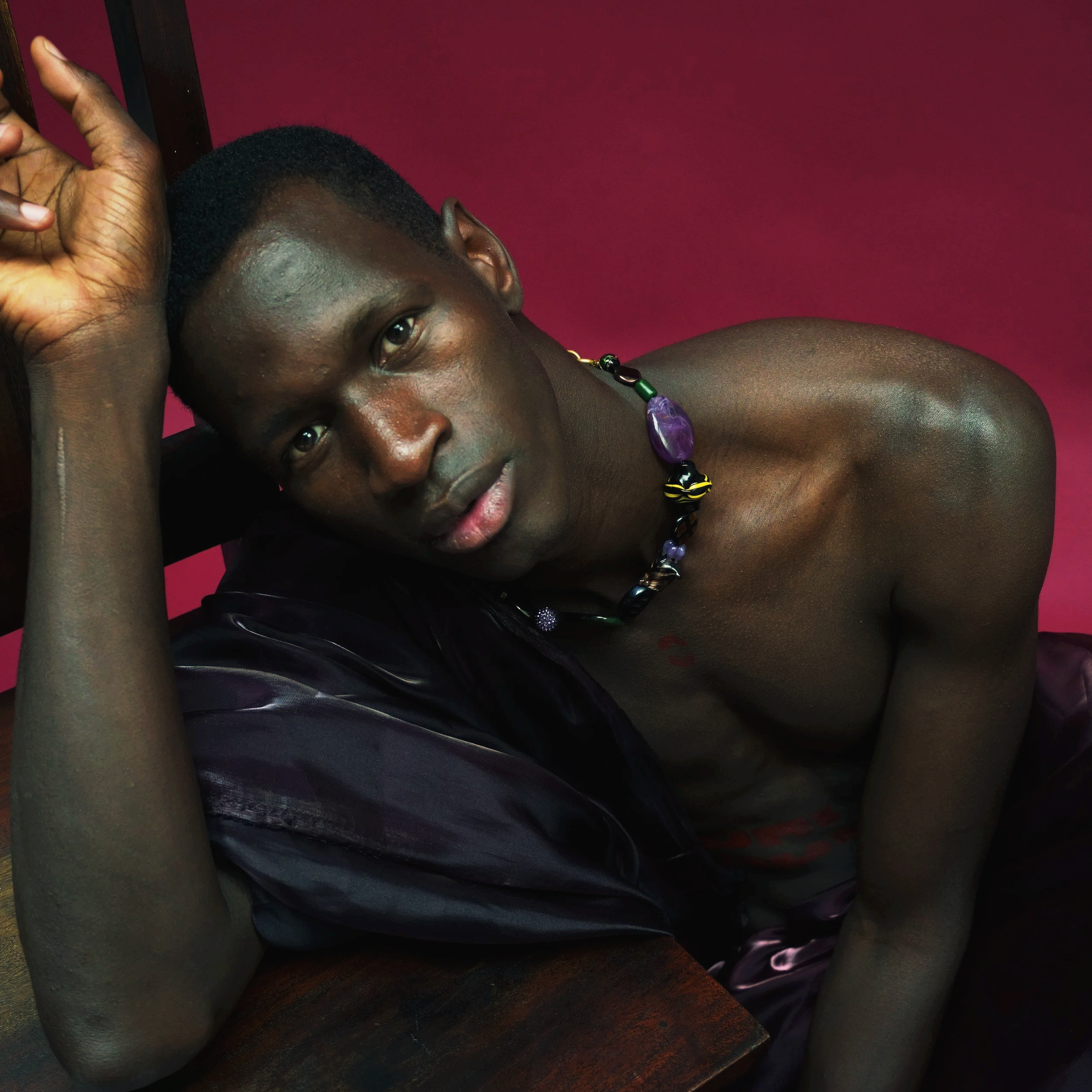 Jeune homme noir allongé, le corps nu, portant un collier en perles colorées, reposant sur une surface en bois avec un fond rose