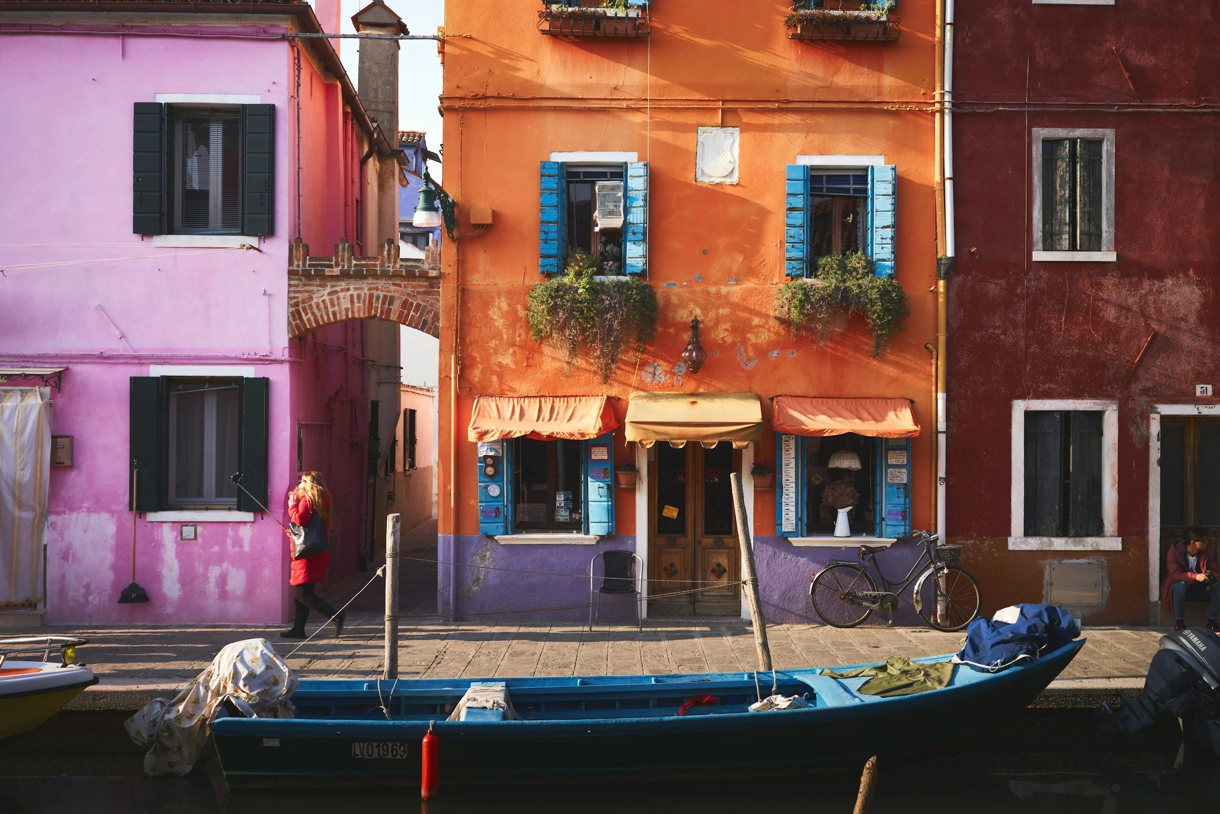 Maisons colorées en Italie avec une façade orange, rose et rouge, fenêtres avec volets bleus et plantes en balcon, un bateau et une personne à côté de l'eau.
