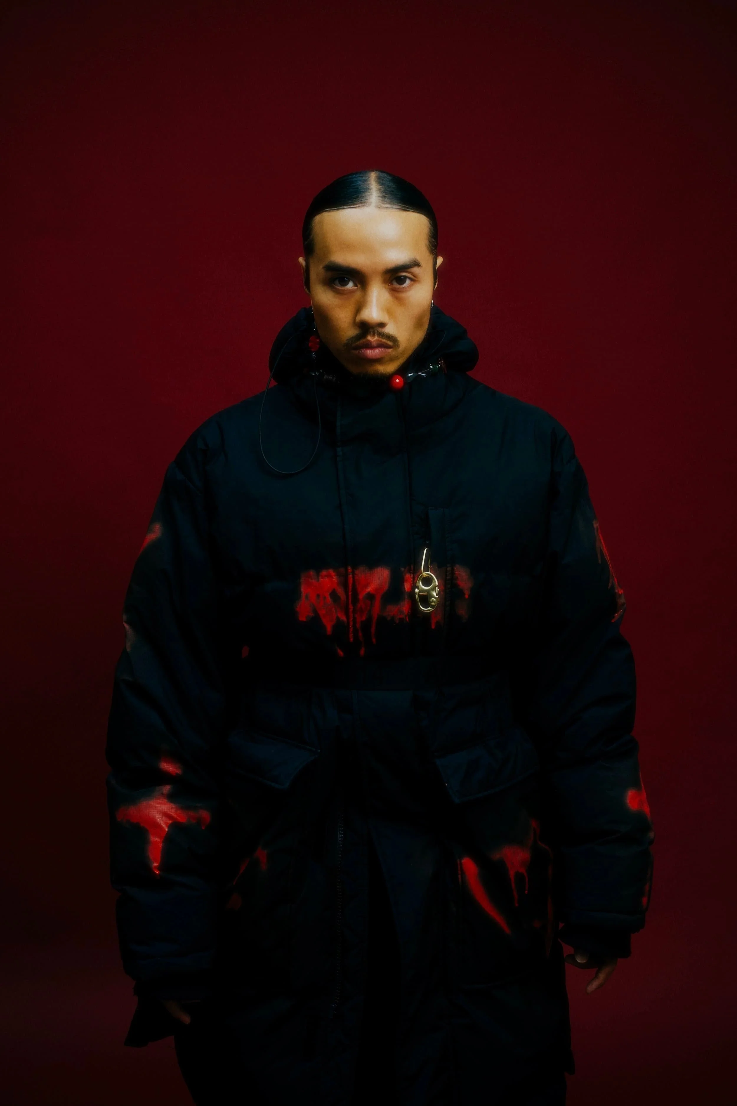 Un homme avec des cheveux noirs attachés, portant une veste noire avec des éclaboussures rouges, devant un fond rouge foncé.