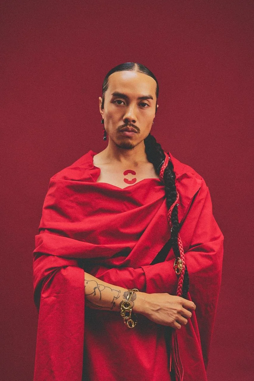 Une personne au regard sérieux portant une robe rouge avec un foulard assorti, des tatouages visibles sur le bras, des boucles d'oreilles longues, et une chevelure noire tressée, posant devant un fond rouge.