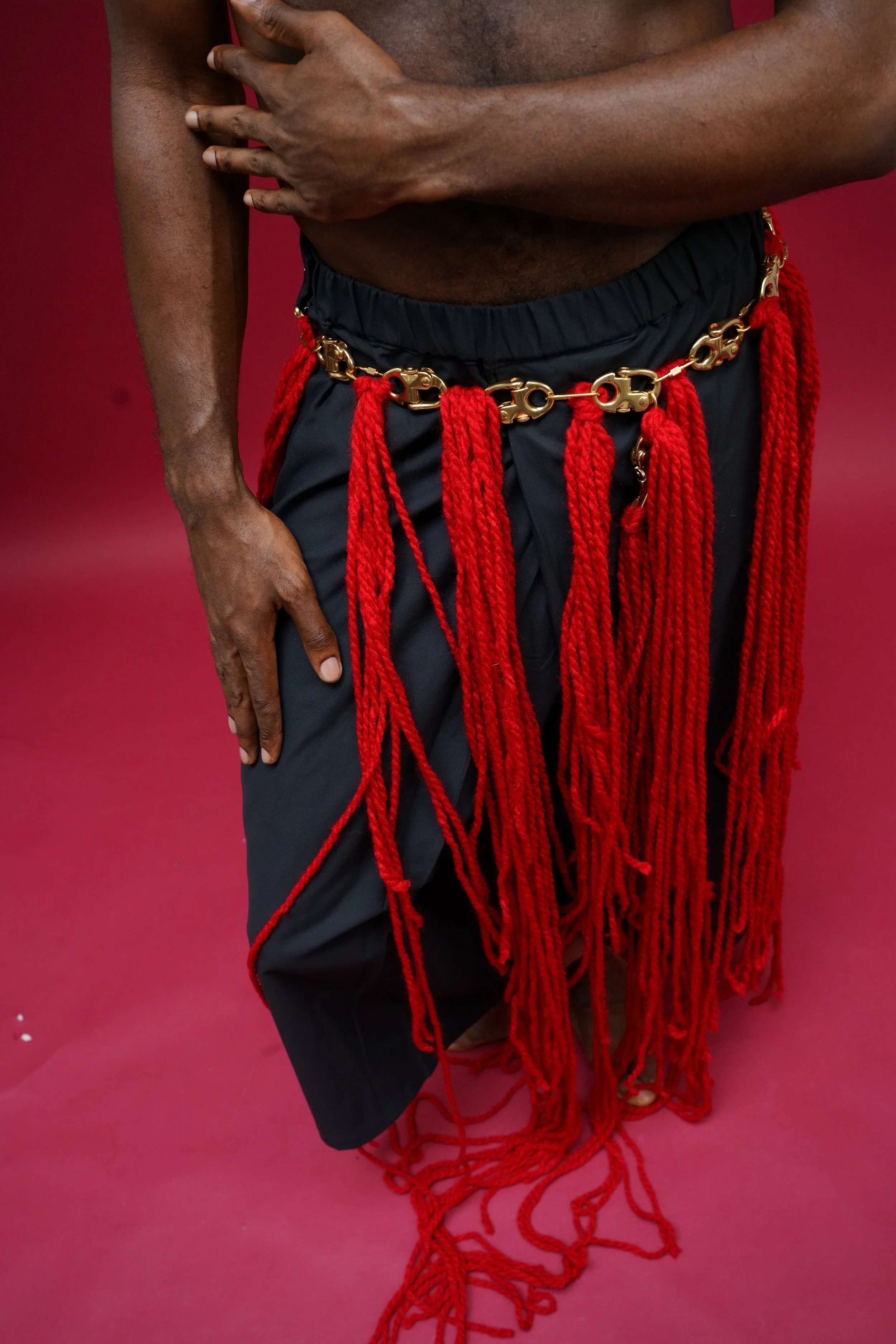 Une personne portant une jupe ou un pantalon noir avec des cordons rouges en chaîne et en fil, devant un fond rouge.