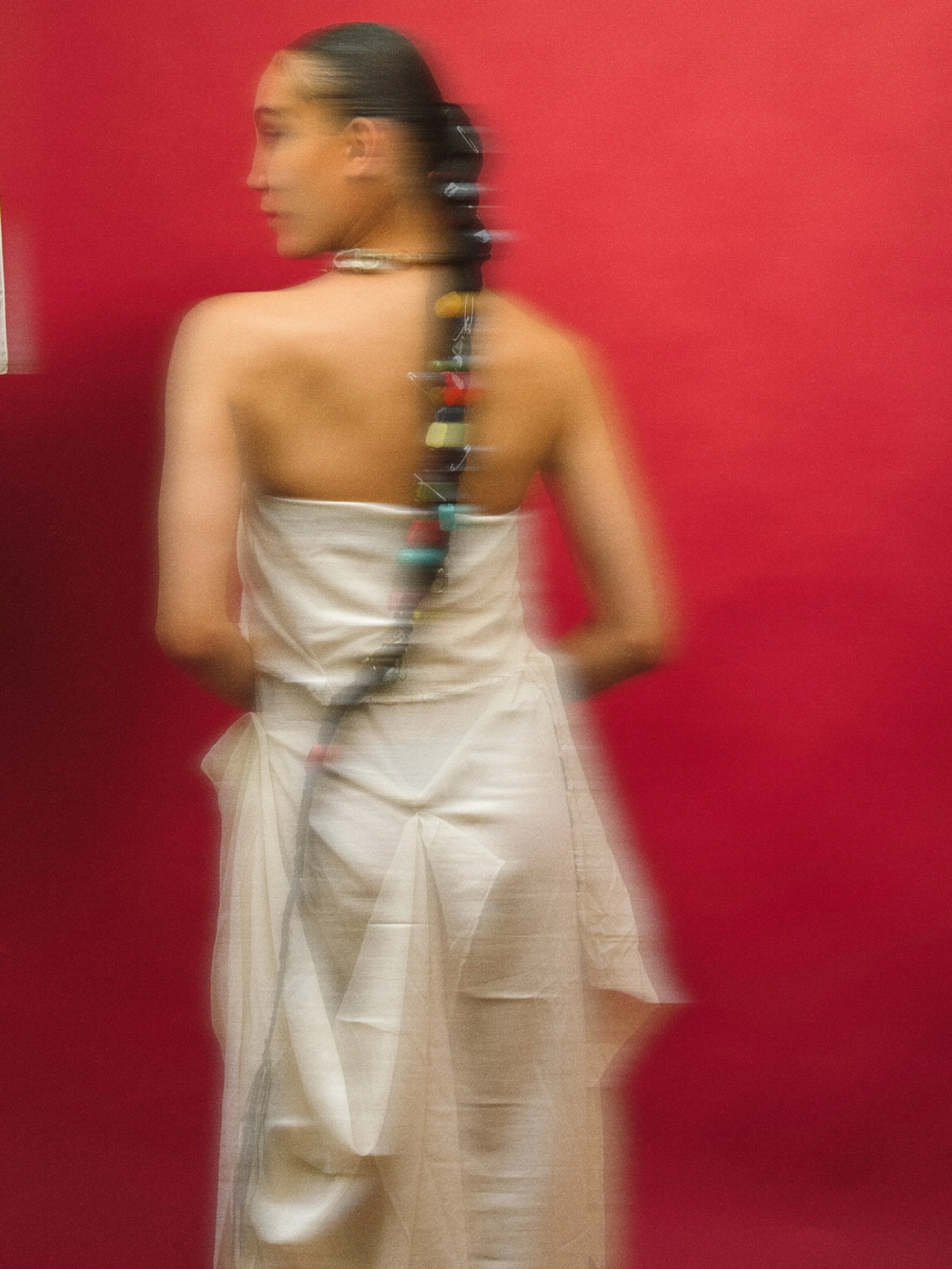 Une femme avec une longue tresse colorée, portant un drapé blanc, debout devant un mur rouge.