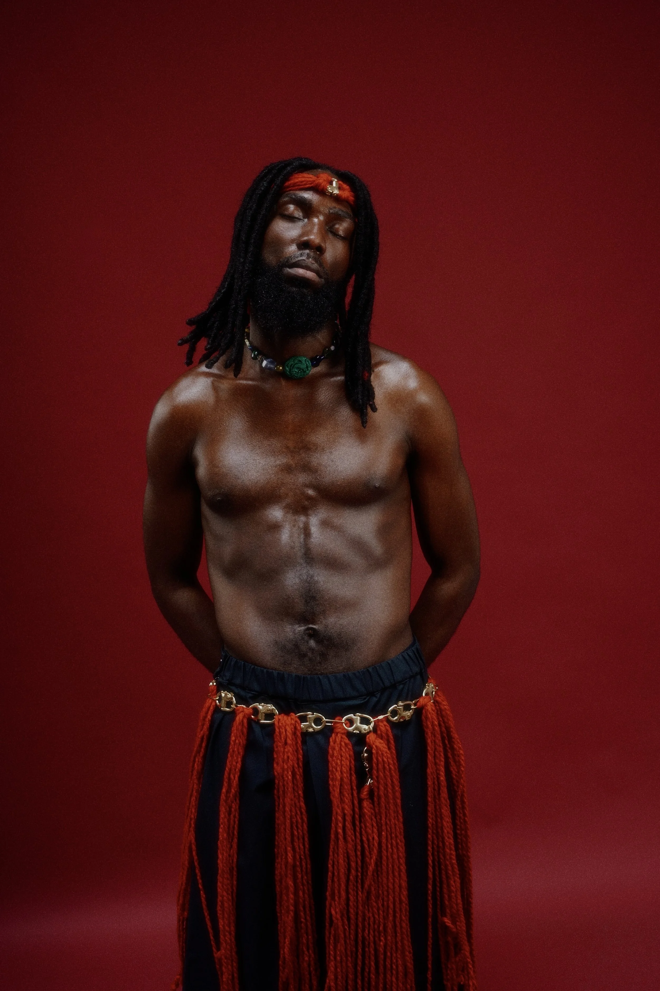 Homme nu portant un collier et une ceinture en chaîne avec des franges rouges, se tenant contre un fond rouge, avec une expression de méditation ou de sérénité.